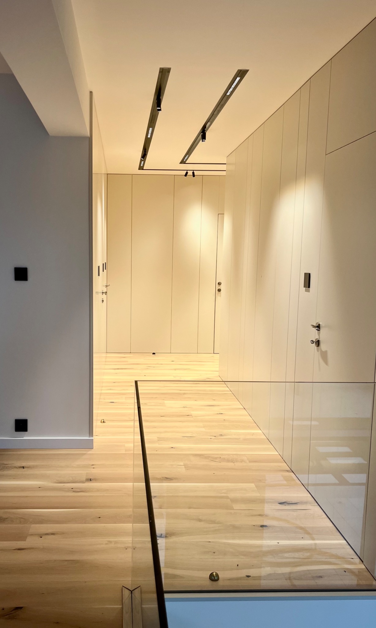 Minimalistyczny korytarz z drewnianą podłogą, białymi ścianami z ukrytymi drzwiami i nowoczesnym oświetleniem szynowym.