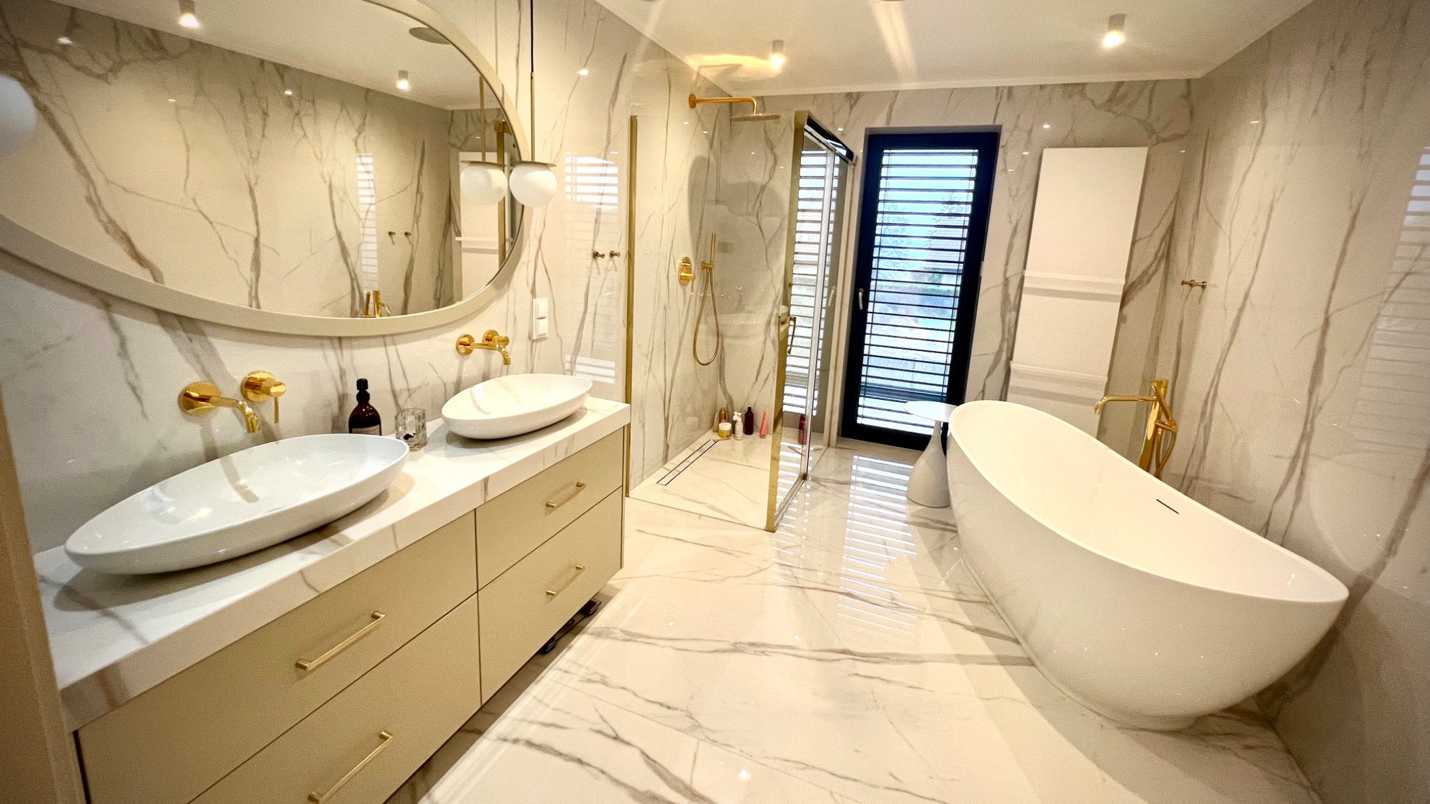 Elegancka łazienka z marmurowymi ścianami i podłogą, wyposażona w dwie białe umywalki nablatowe z złotymi bateriami, owalne lustro z oświetleniem, kabinę prysznicową ze złotymi detalami...