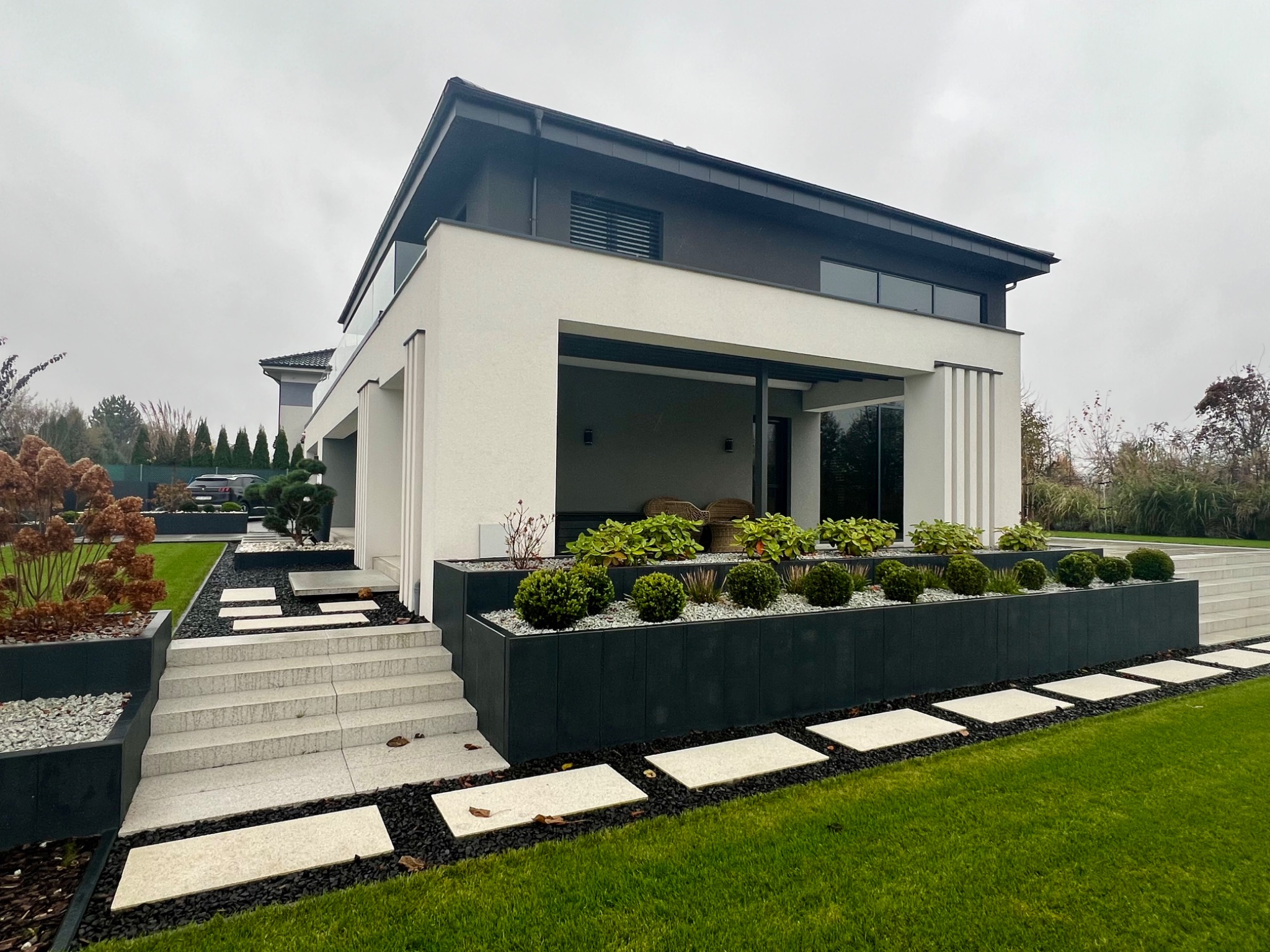 Nowoczesny dwukondygnacyjny dom z płaskim dachem i ogrodem z geometrycznymi rabatami, jasnymi płytami chodnikowymi i ciemnym żwirem, widok z niskiego kąta na trawnik.