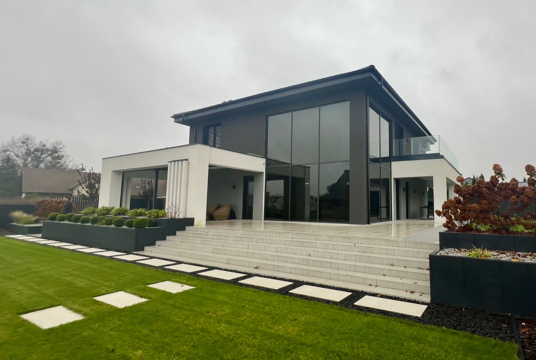 Nowoczesny dom z dużymi przeszkleniami, schodami wejściowymi i zadbanym trawnikiem z kamiennymi płytami.