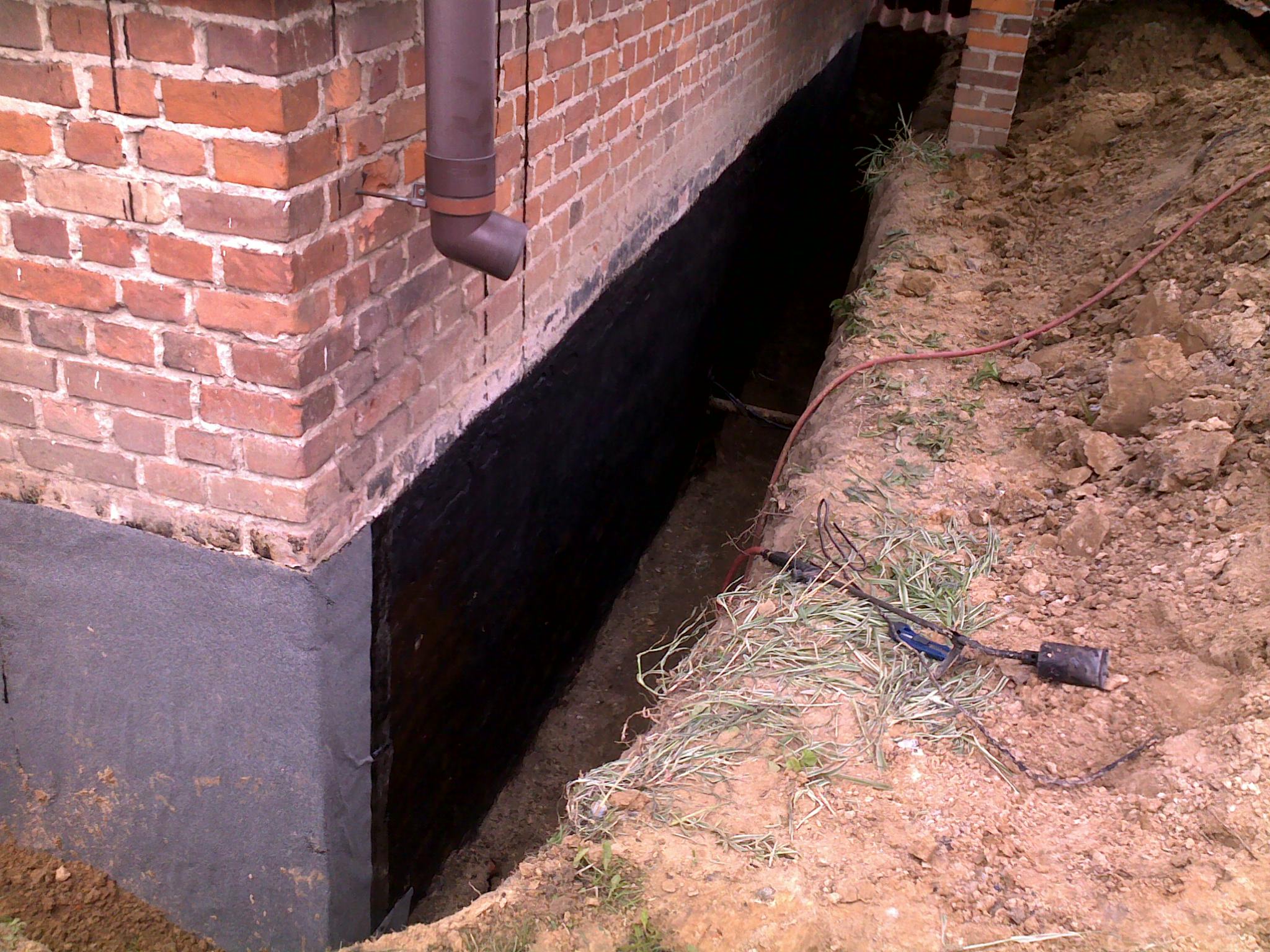 Izolacja pionowa fundamentów z cegły, zabezpieczona czarną powłoką hydroizolacyjną, widoczny wykop w gruncie.