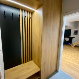 Shakira Monika Mathea - Zabudowa przedpokoju z szafą, siedziskiem i panelem ściennym z wieszakami, oświetlona listwą LED, widok na wnętrze domu z drewnianą podłogą.