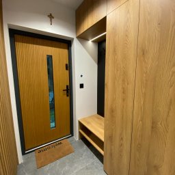 Shakira Monika Mathea - Nowoczesny przedpokój z zabudowaną szafą z drewnianymi frontami, siedziskiem i wieszakami, widoczna również drewniana drzwi z czarną ramą i małym okienkiem oraz wycieraczka z napisem 'welcome'.