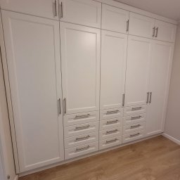 Shakira Monika Mathea - Biała szafa wnękowa z szufladami i górnymi szafkami, srebrne uchwyty, panele podłogowe imitujące drewno.