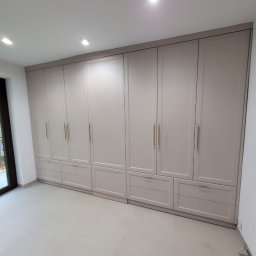 Shakira Monika Mathea - Jasnoszara szafa na wymiar z złotymi uchwytami, zabudowa od podłogi do sufitu, widok z przodu na tle jasnej podłogi i ściany, minimalistyczny design.