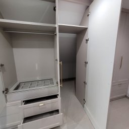 Shakira Monika Mathea - Otwarta szafa wnękowa z szufladami i półkami, widoczna przestrzeń garderoby za nią. Jasne kolory, minimalistyczny design, złote uchwyty. Widok z bliska.