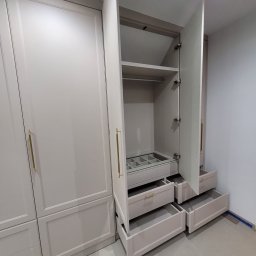 Shakira Monika Mathea - Jasna szafa wnękowa z otwartymi drzwiami i szufladami, ukazująca wewnętrzną organizację przestrzeni. Widoczne półki, szuflady z przegródkami i złote uchwyty.