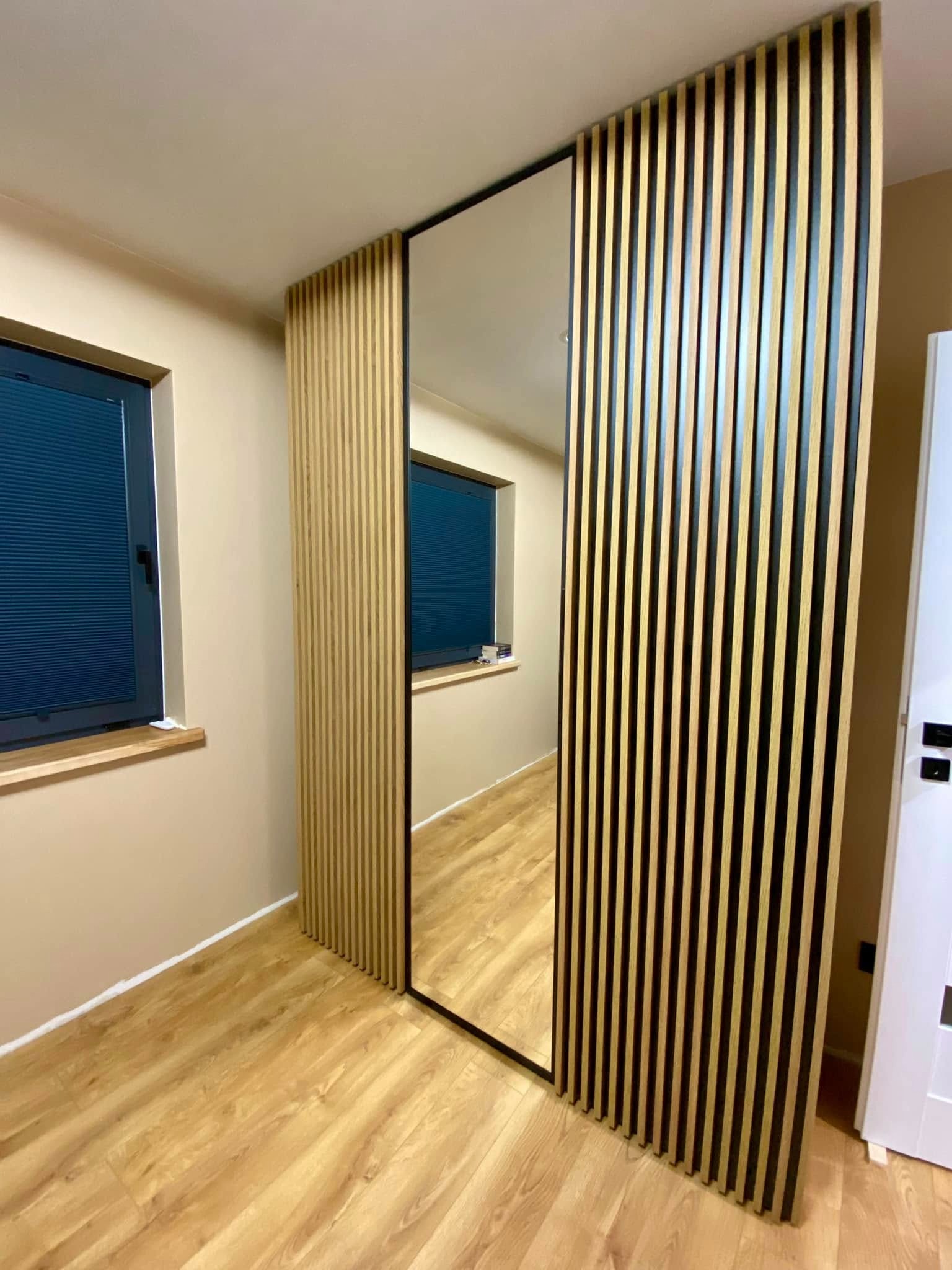 Nowoczesna szafa wnękowa z lustrem i lamelami w odcieniach drewna i czerni, tworząca przegrodę w jasnym wnętrzu z drewnianą podłogą i oknem z żaluzjami.