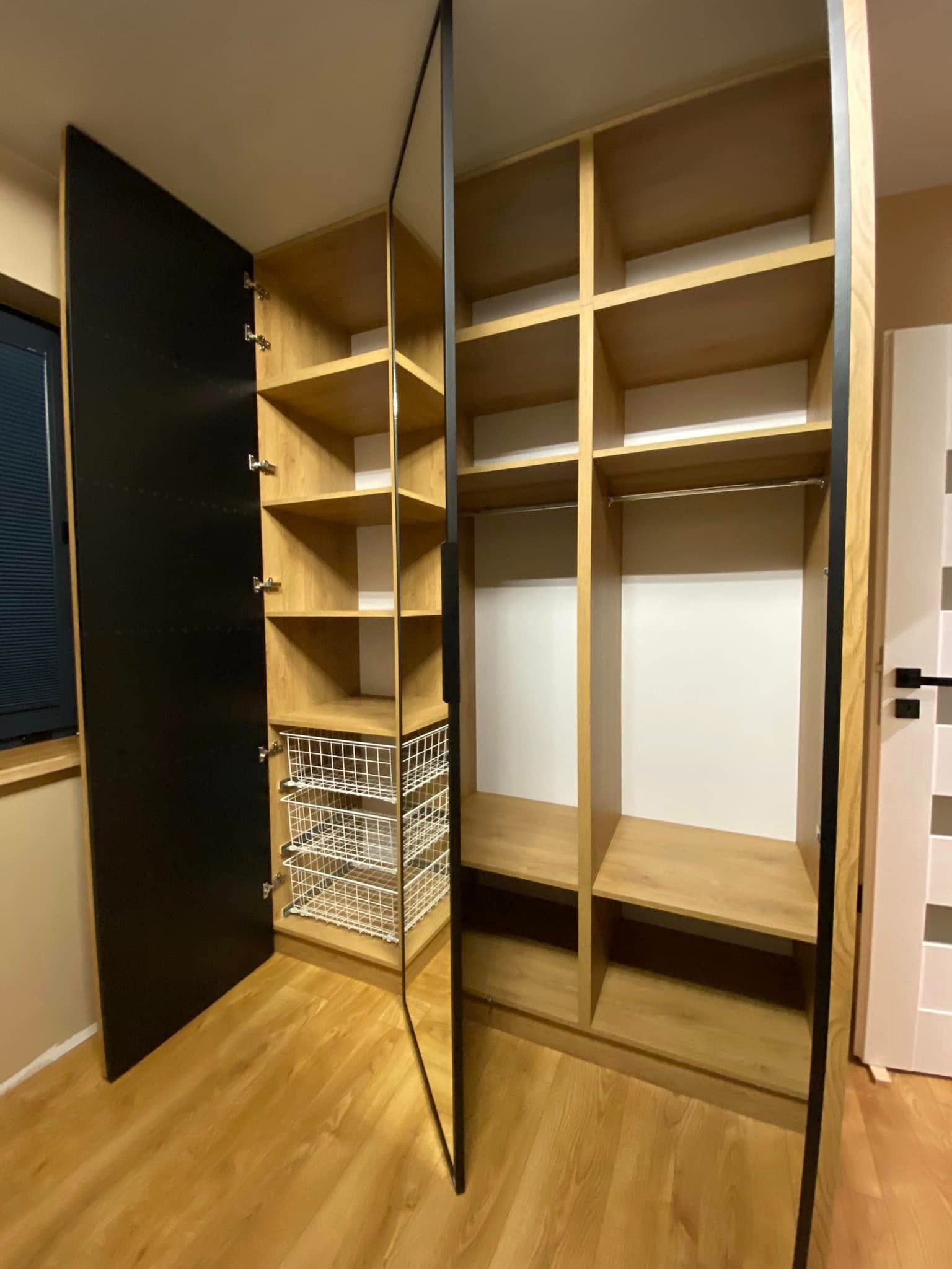 Otwarta szafa wnękowa z drewnianymi półkami i czarnym frontem, widoczne metalowe kosze na prowadnicach. Część szafy z drążkiem na ubrania i białym wnętrzem. Na podłodze panele imitujące drewno.