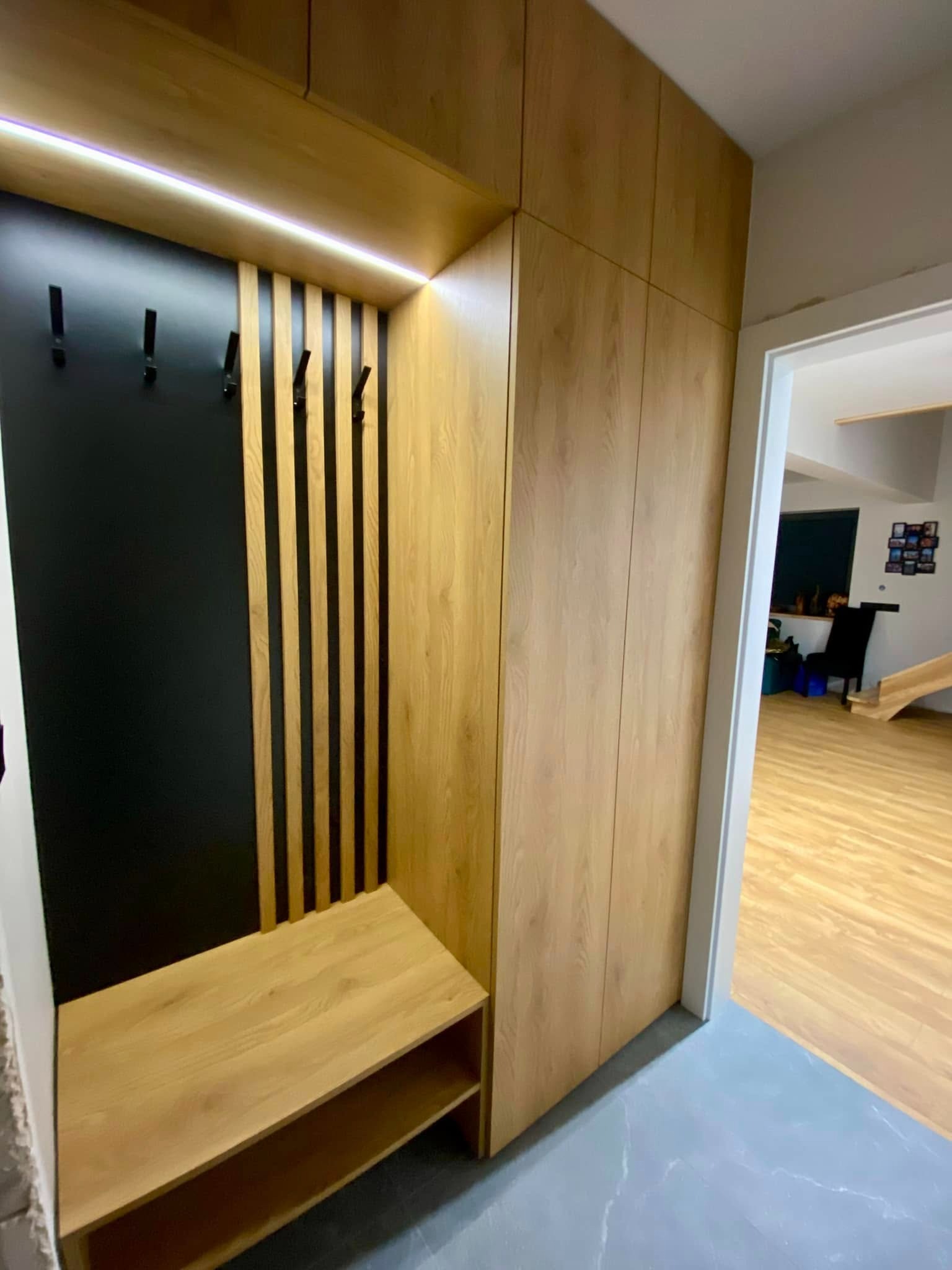 Zabudowa przedpokoju z szafą, siedziskiem i panelem ściennym z wieszakami, oświetlona listwą LED, widok na wnętrze domu z drewnianą podłogą.