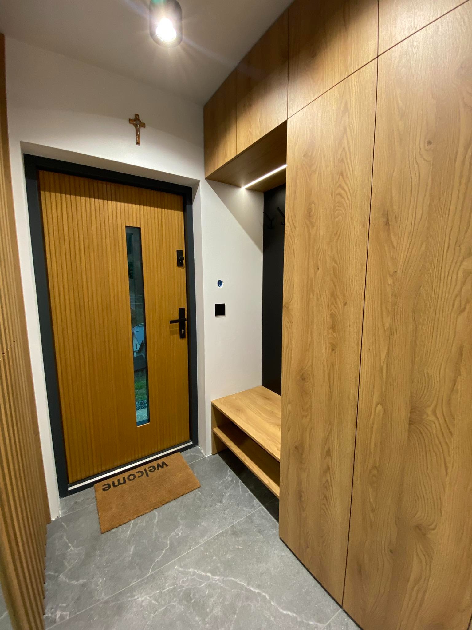 Nowoczesny przedpokój z zabudowaną szafą z drewnianymi frontami, siedziskiem i wieszakami, widoczna również drewniana drzwi z czarną ramą i małym okienkiem oraz wycieraczka z napisem 'welcome'.