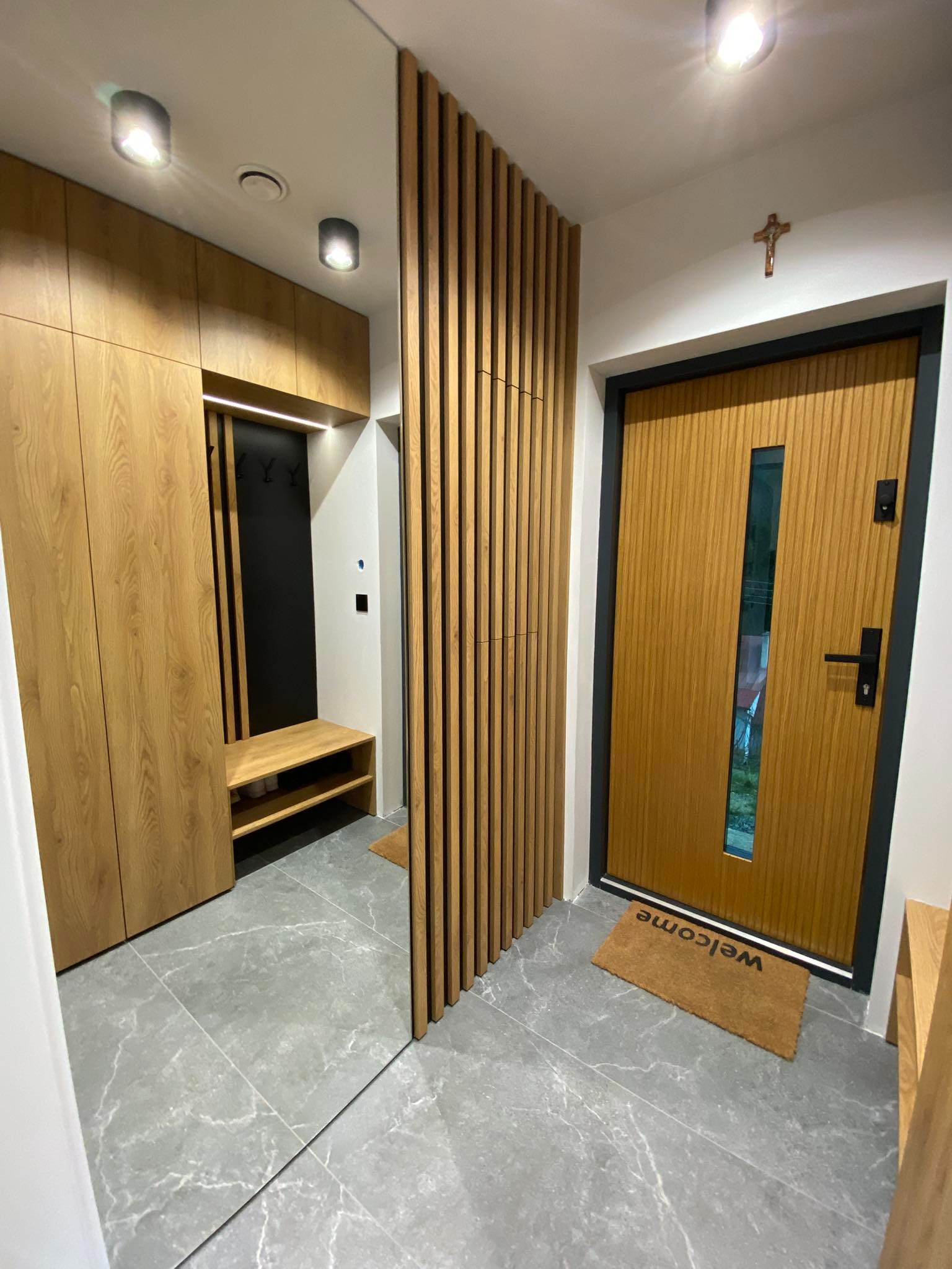 Nowoczesny przedpokój z drewnianą zabudową, lustrem i lamelami, szara podłoga z płytek imitujących marmur, brązowa wycieraczka przed drzwiami wejściowymi.