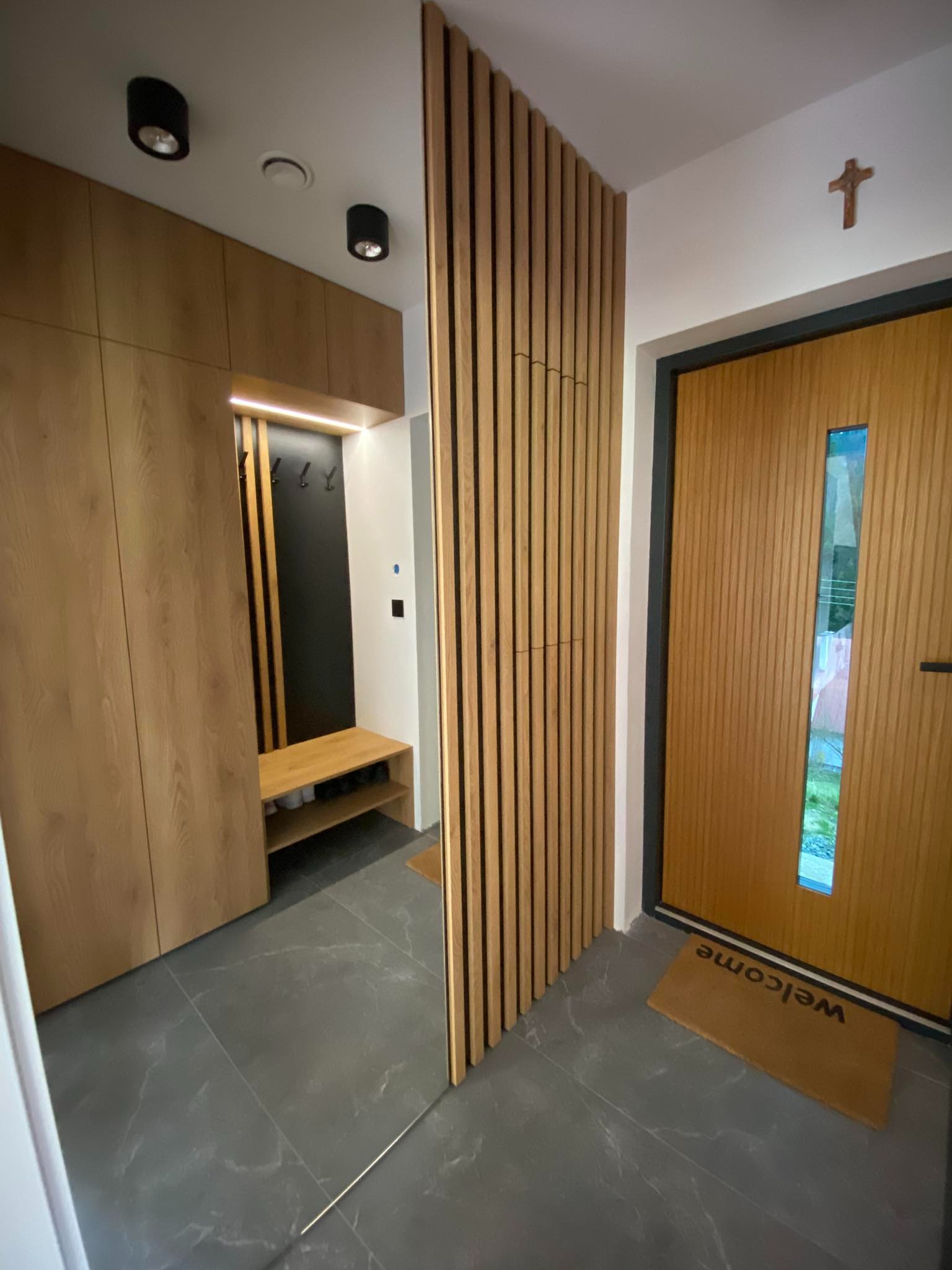 Nowoczesny przedpokój z zabudową z drewnianych paneli i szafą z jasnego drewna, uzupełniony o minimalistyczne oświetlenie sufitowe i drewniane drzwi wejściowe z przeszkleniem.