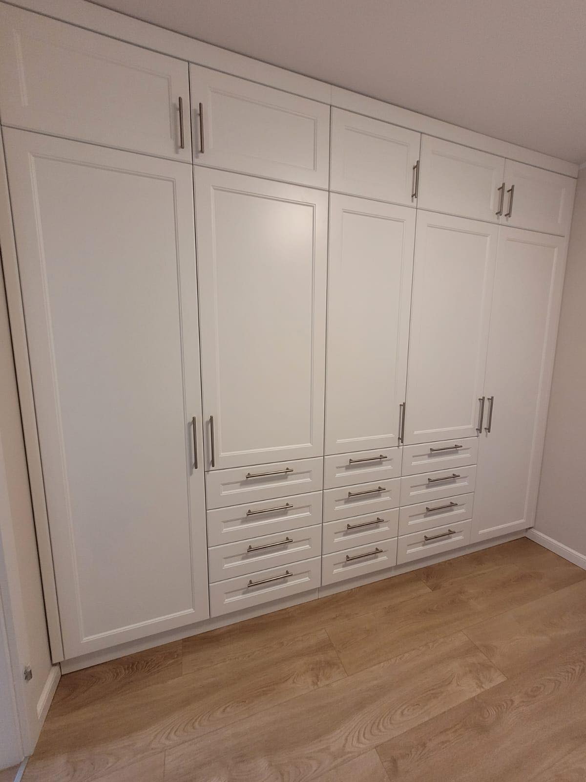 Biała szafa wnękowa z szufladami i górnymi szafkami, srebrne uchwyty, panele podłogowe imitujące drewno.