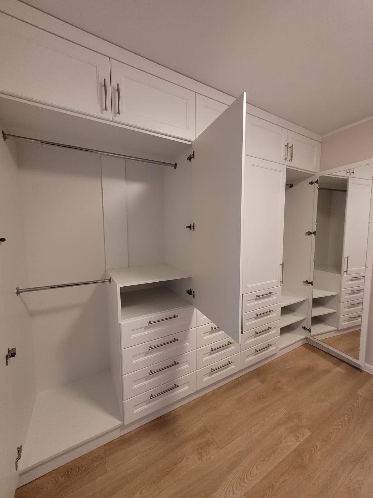 Biała szafa wnękowa z otwartymi drzwiami, widoczne półki, szuflady i drążek na ubrania, lustro na jednym z frontów odbija wnętrze szafy