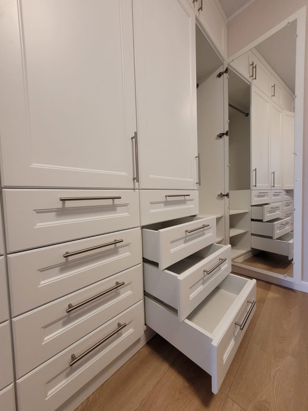 Biała szafa wnękowa z szufladami i lustrzanymi drzwiami, trzy szuflady wysunięte, widoczne półki wewnątrz szafy.