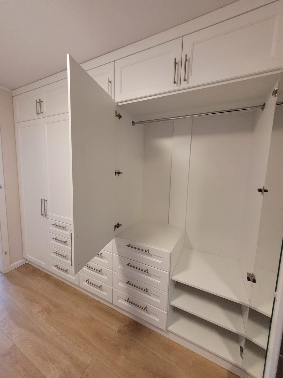 Biała szafa wnękowa z otwartymi drzwiami, ukazująca półki i szuflady z metalowymi uchwytami, drewniana podłoga.