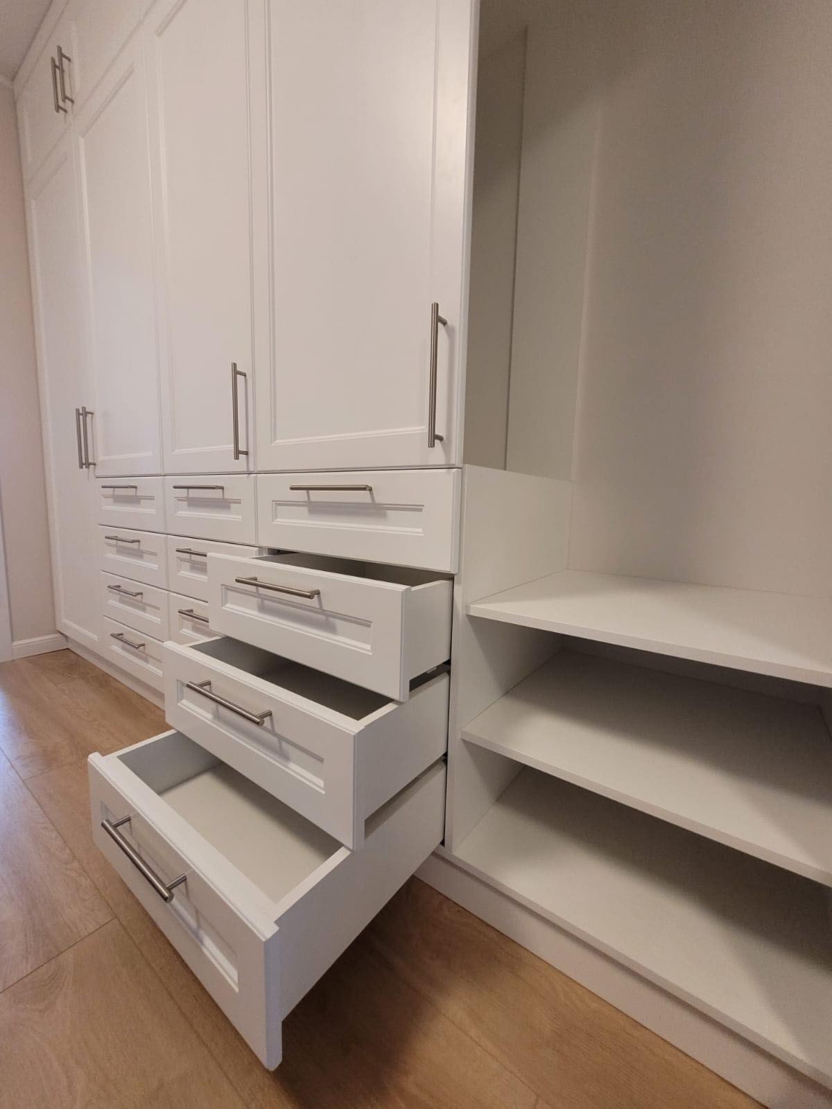 Biała szafa wnękowa z otwartymi szufladami i półkami, widok z dołu, drewniana podłoga.