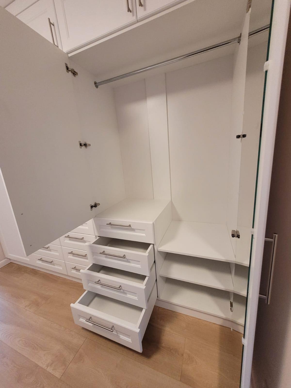 Wnętrze białej szafy wnękowej z otwartymi szufladami i półkami, widoczne okucia i reling na ubrania.