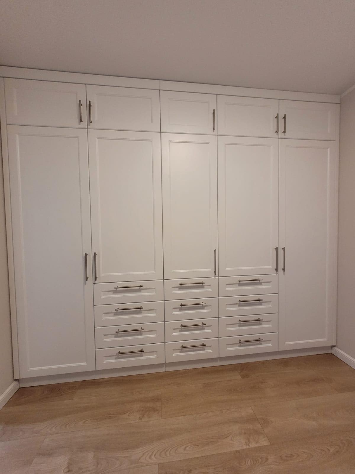 Biała szafa wnękowa z górnymi szafkami i rzędem szuflad, srebrne uchwyty, panele podłogowe w kolorze jasnego drewna.