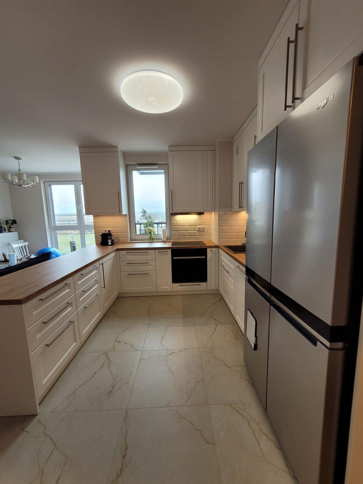 Jasna kuchnia w stylu angielskim z drewnianym blatem, białymi szafkami i płytkami imitującymi cegłę, oświetlona lampą sufitową z efektem gwiazd.