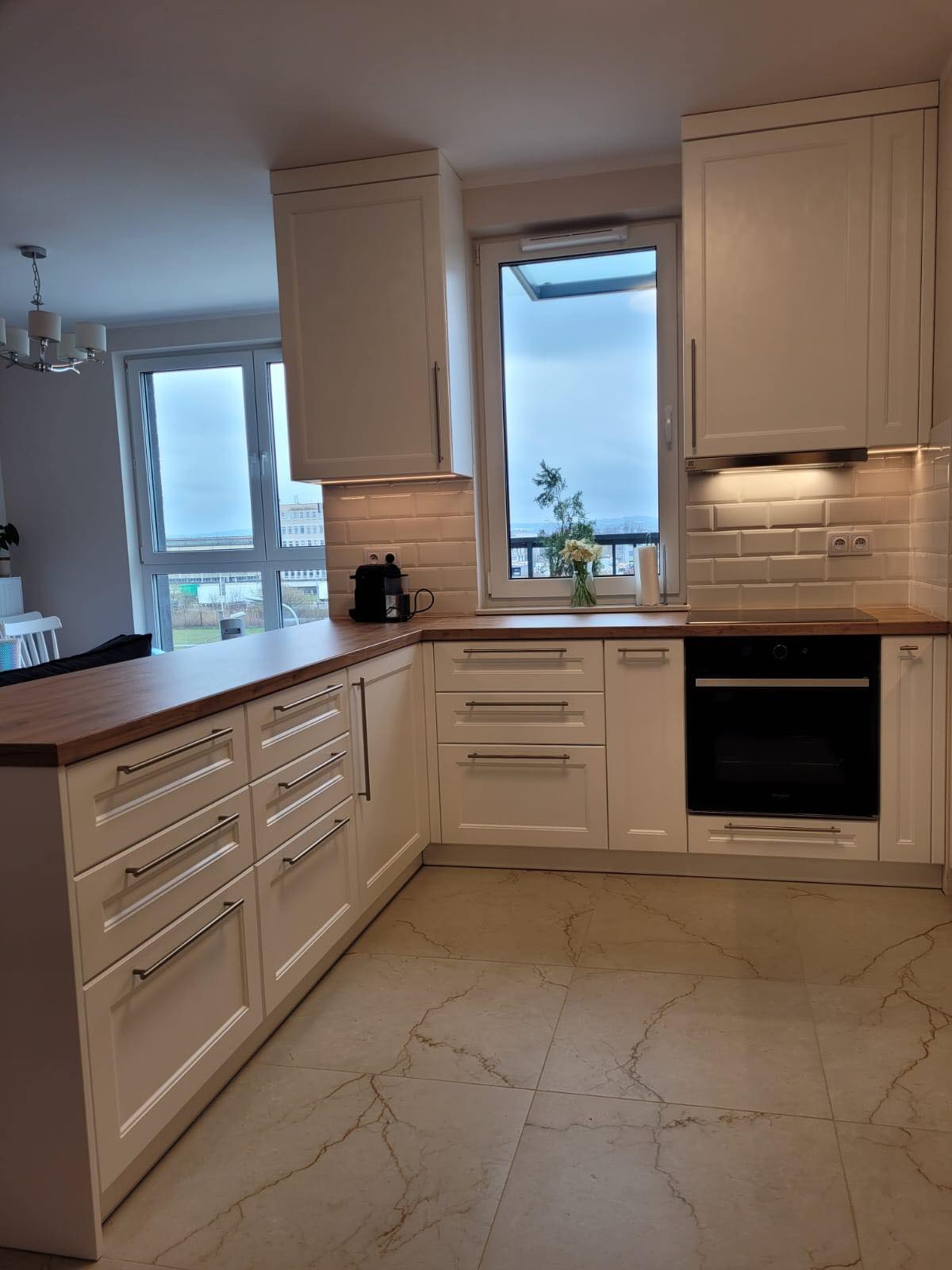 Jasna kuchnia w stylu angielskim z białymi szafkami, drewnianym blatem i widokiem na miasto zza okna, z ekspressem do kawy i kwiatami na blacie.