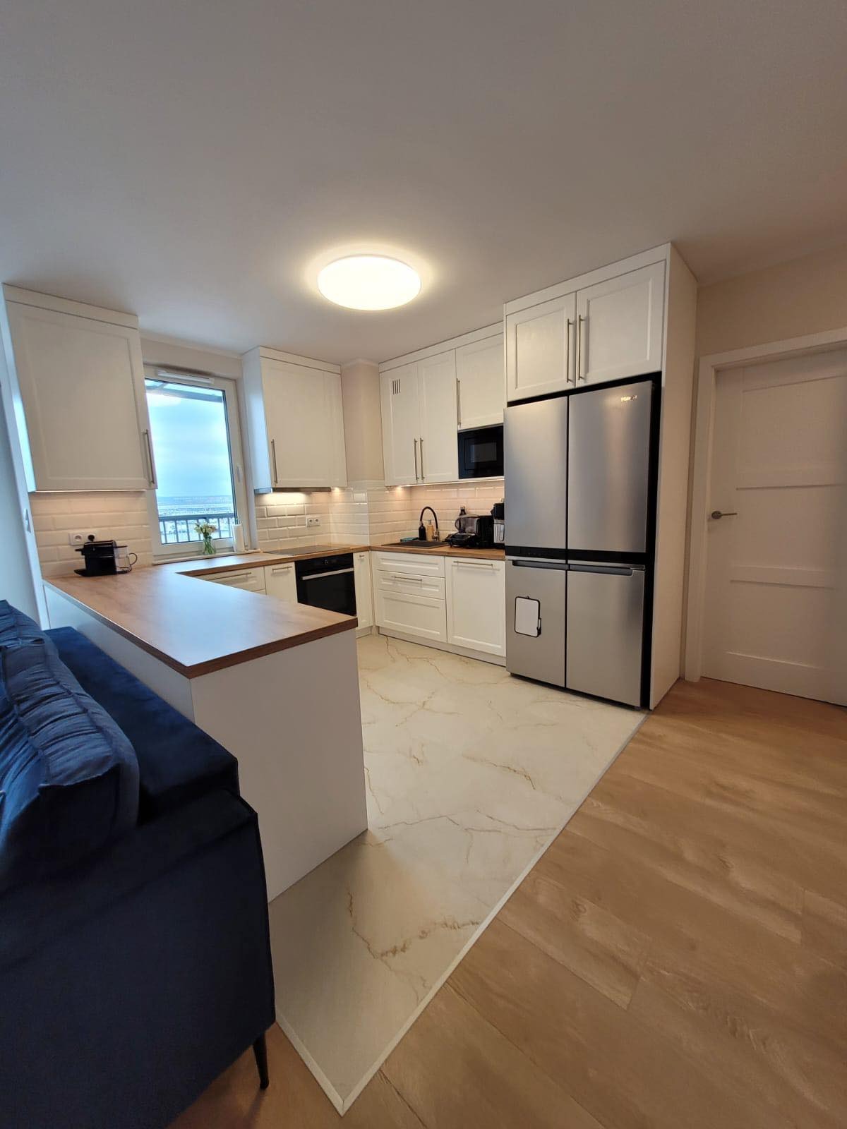 Jasna kuchnia z białymi szafkami, drewnianym blatem i dużym, srebrnym lodówko-zamrażarką; widok z salonu na okno z widokiem na morze.