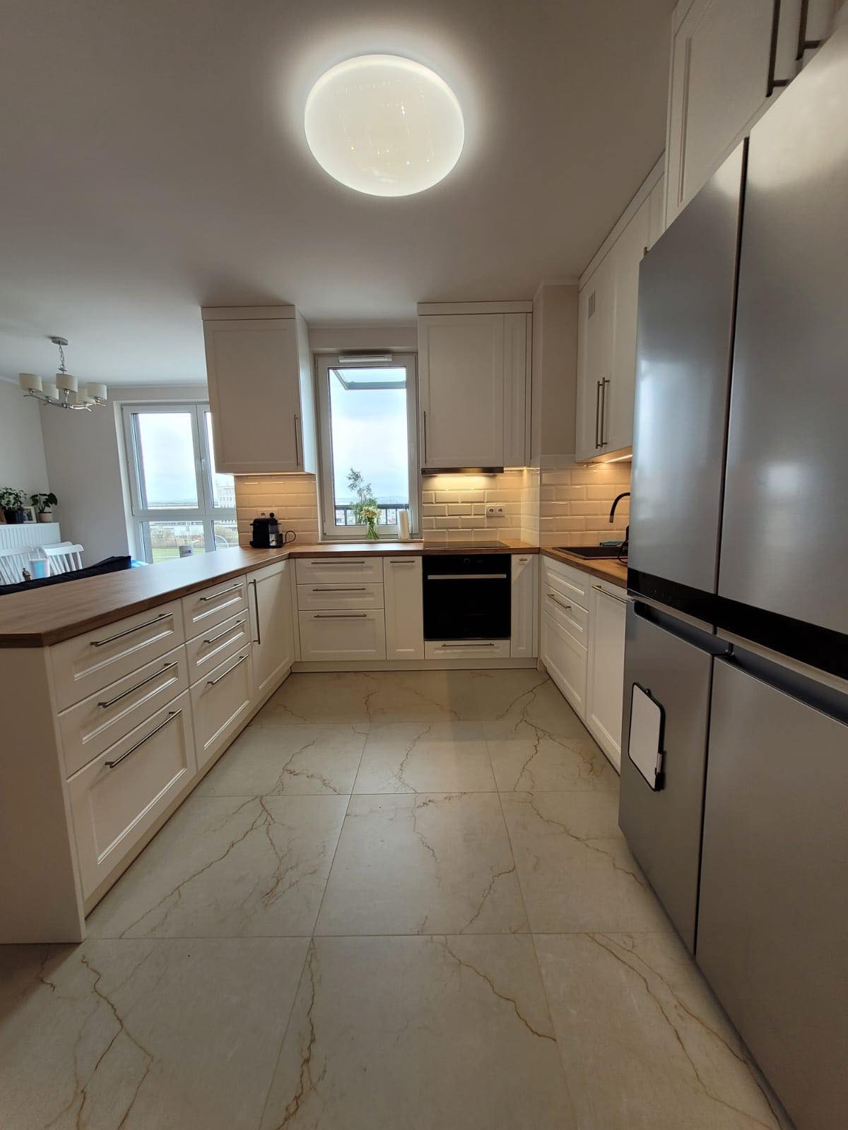 Jasna kuchnia w stylu angielskim z białymi frontami, drewnianym blatem i marmurową podłogą, oświetlona światłem dziennym i lampkami podszafkowymi.