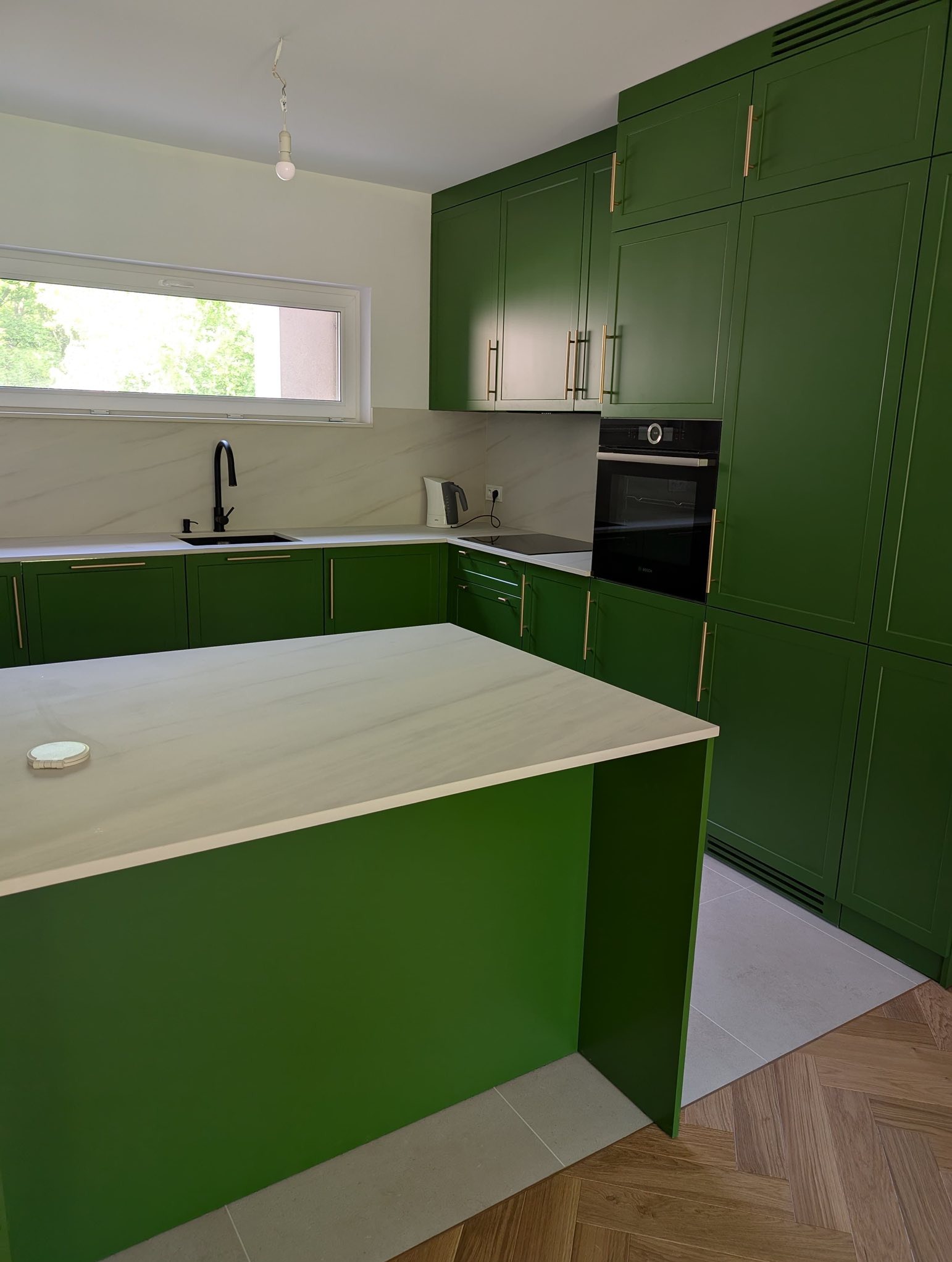 Kuchnia w stylu angielskim: zielone szafki z złotymi uchwytami, biały blat i marmurowe wykończenie ściany. Widoczny fragment wyspy kuchennej i sprzęt AGD.