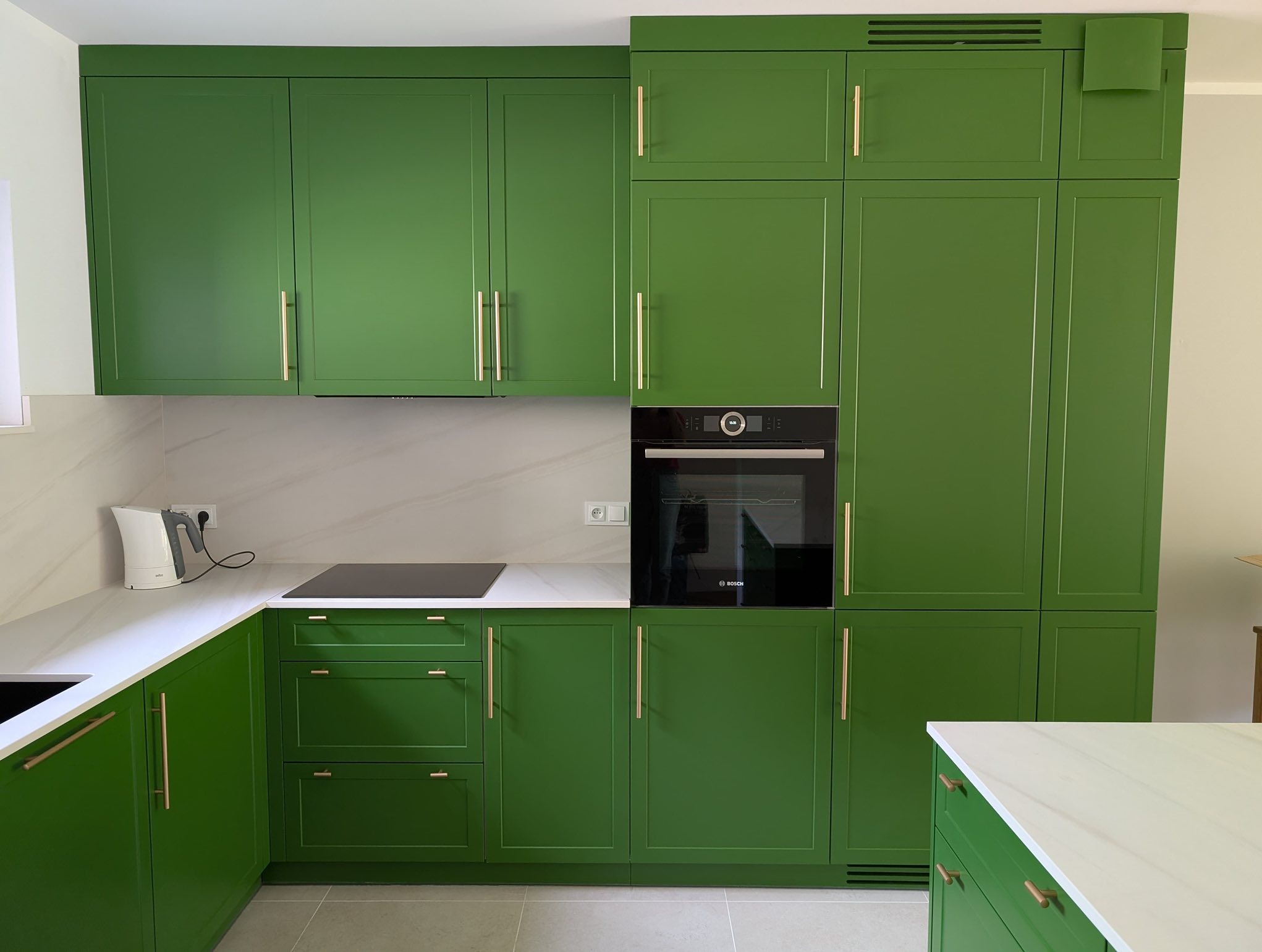 Kuchnia w stylu angielskim, fronty w odcieniu zieleni paproci. Złote, podłużne uchwyty. Płyta indukcyjna i piekarnik w zabudowie. Marmurowy blat i backsplash.