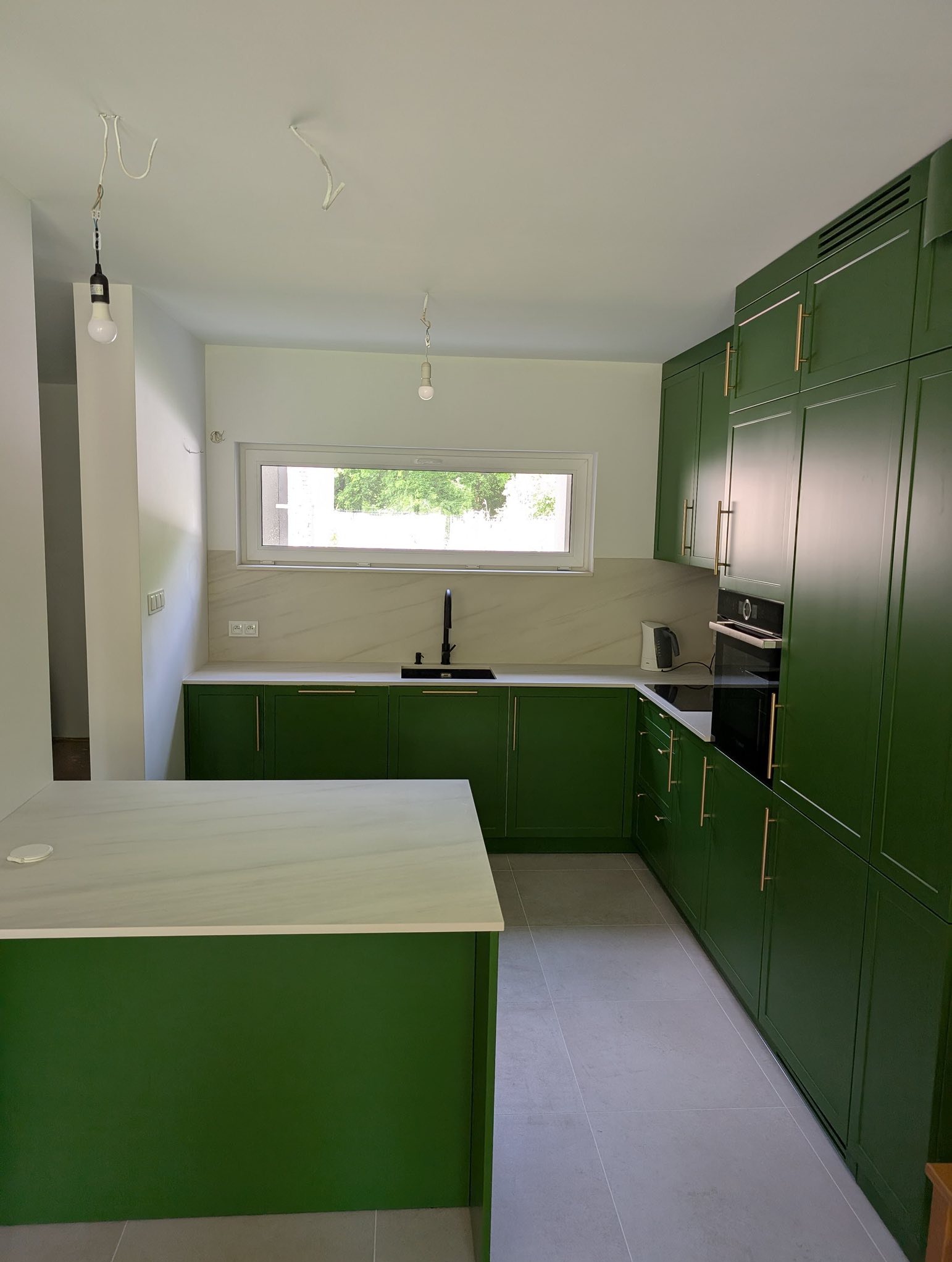 Nowoczesna kuchnia w odcieniach zieleni z marmurowym blatem i złotymi uchwytami. Widoczne okno i minimalistyczne oświetlenie.