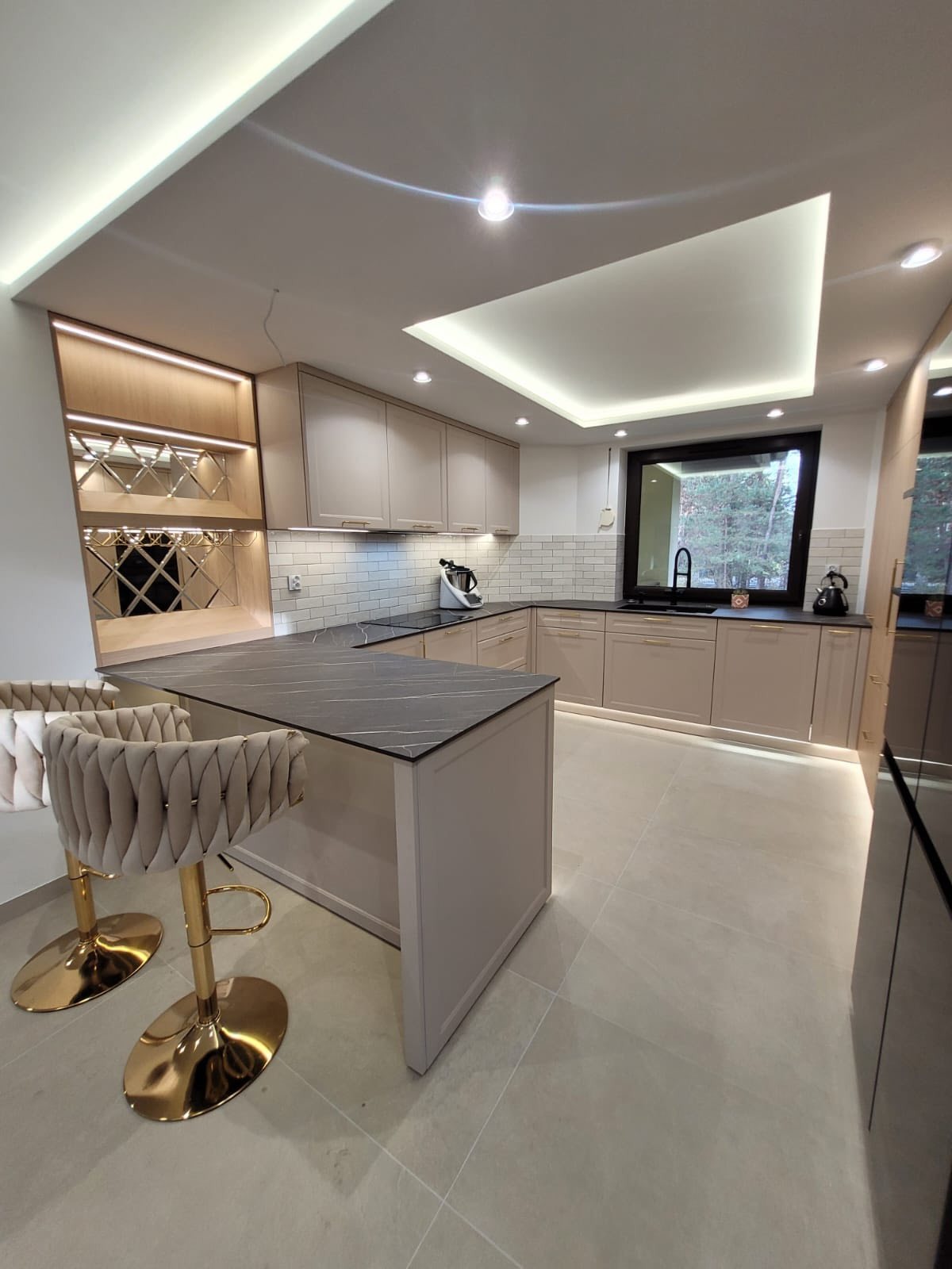 Jasna kuchnia z wyspą i barkiem z regałem na wino. Fronty w kolorze kaszmiru, czarny blat z żyłami, podwieszany zlew i wygodne siedzisko. Oświetlenie LED.