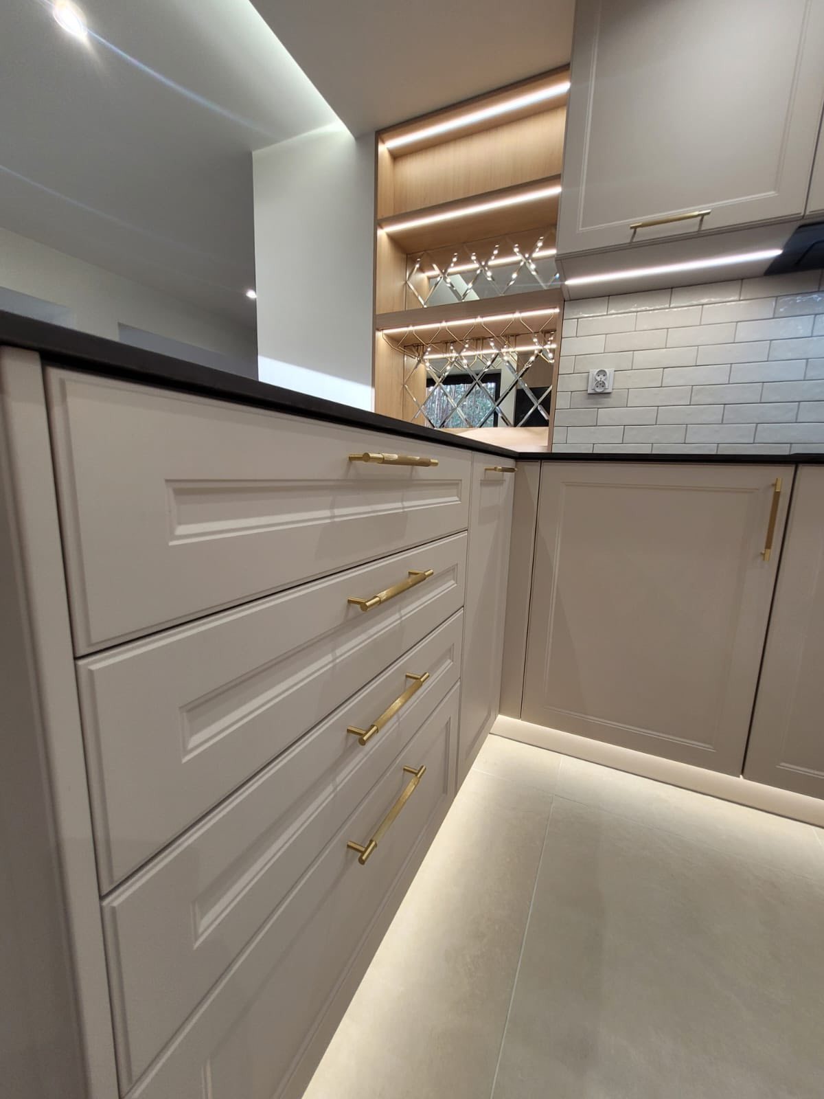 Kremowa zabudowa kuchenna z frezowanymi frontami, złotymi uchwytami i podświetlanym regałem na wino z lustrzanymi plecami. Blat czarny, płytki białe.