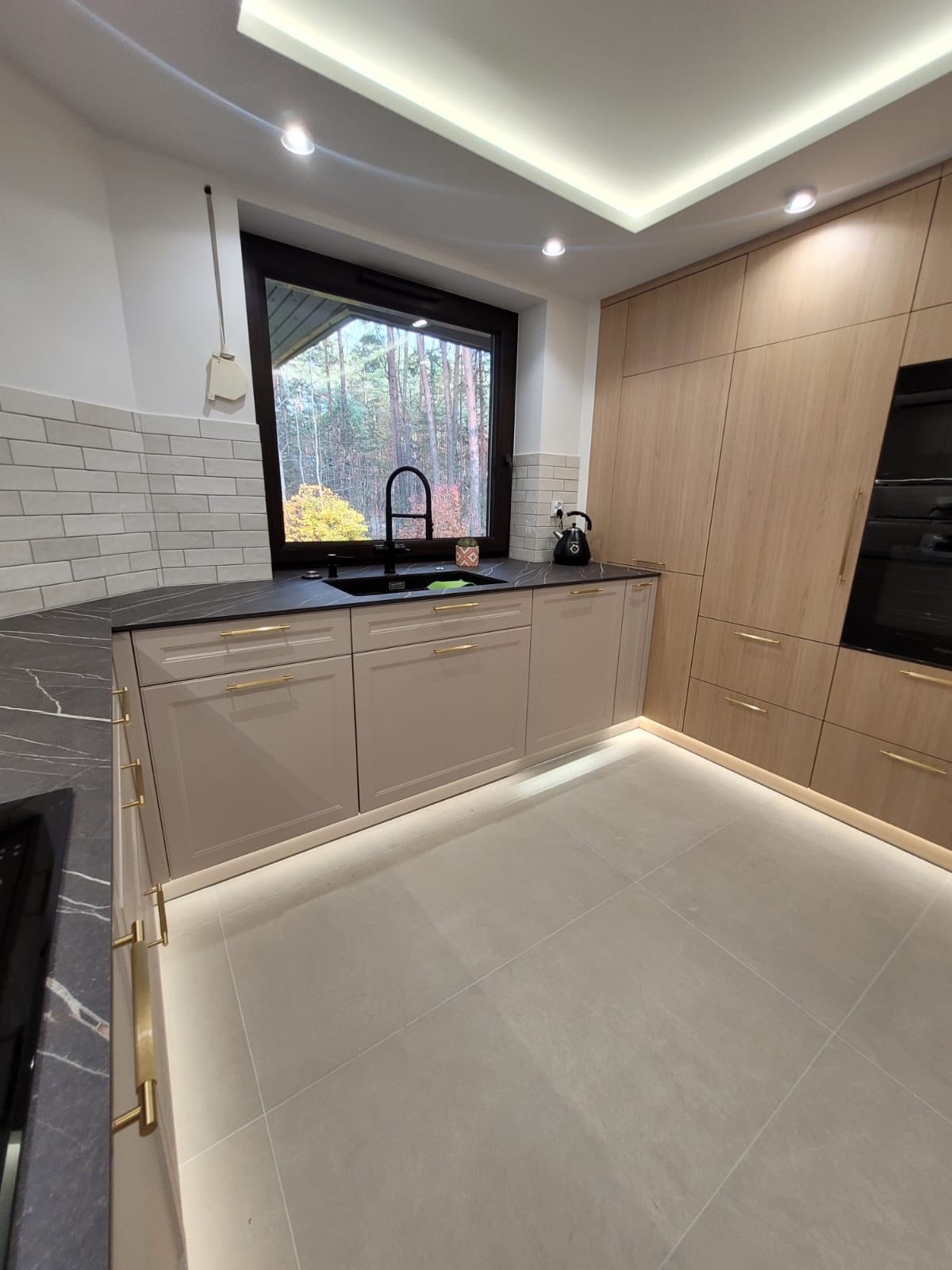 Jasna kuchnia z angielskimi frontami i blatem imitującym marmur. Podświetlenie LED pod szafkami. Za oknem widok na las.