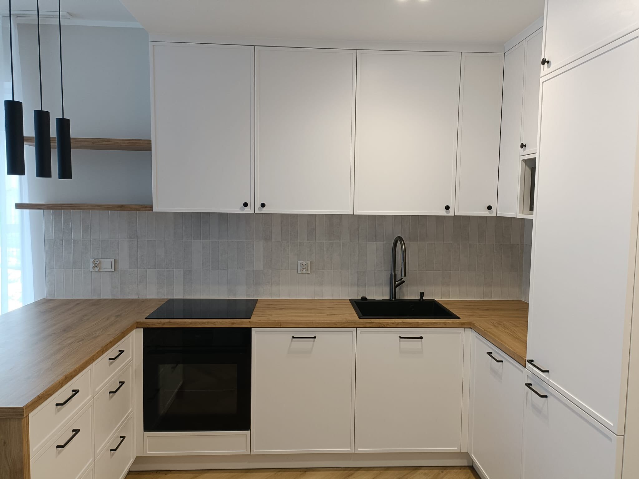 Jasna kuchnia w stylu angielskim: białe, frezowane fronty szafek, drewniany blat, czarne detale (armatura, uchwyty), płytki w odcieniach szarości. Minimalistyczne lampy.