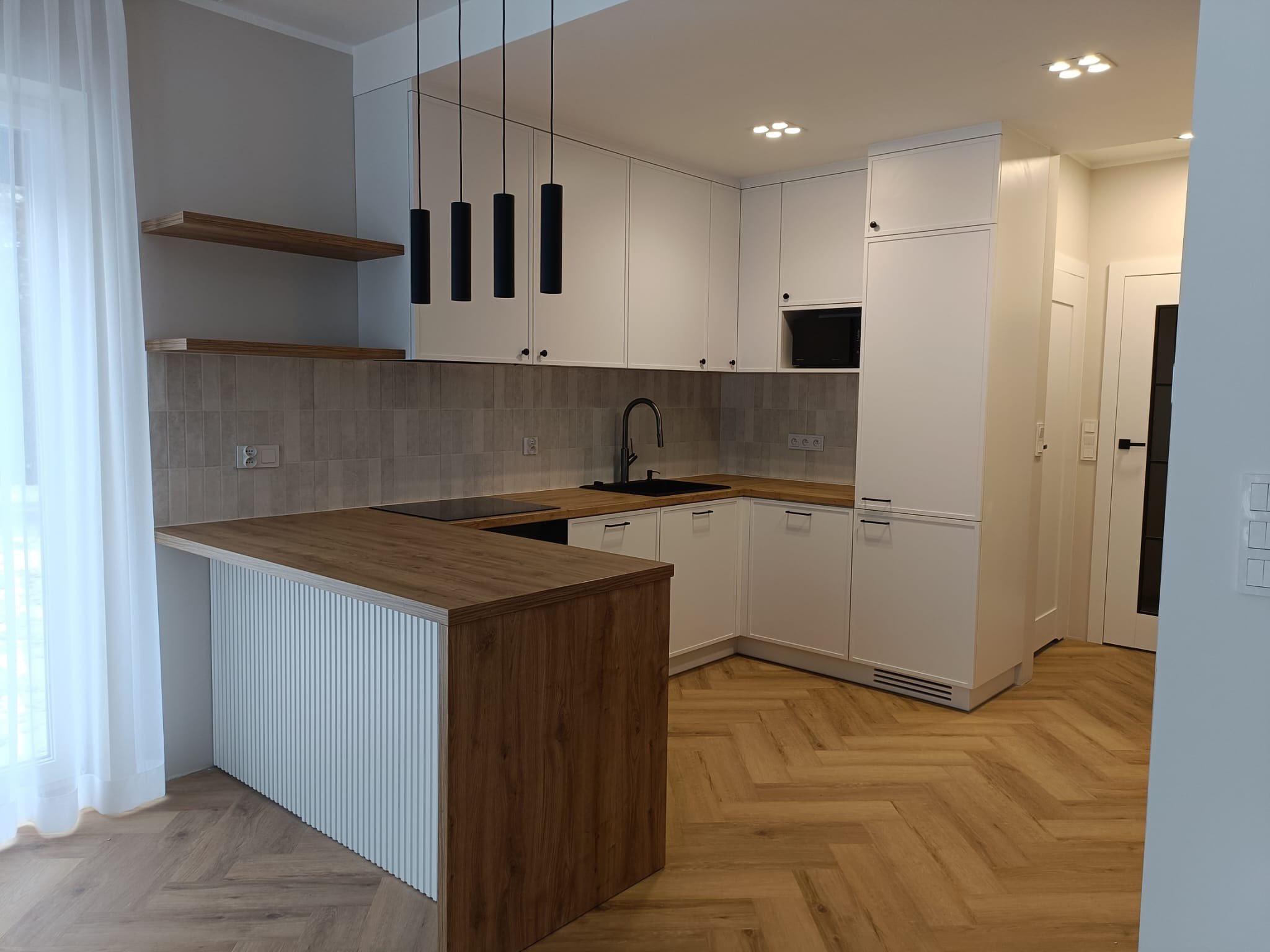 Jasna kuchnia w stylu angielskim z drewnianym blatem i wyspą z ryflowanym frontem. Minimalistyczne czarne lampy wiszące nad wyspą. Parkiet w jodełkę na podłodze.