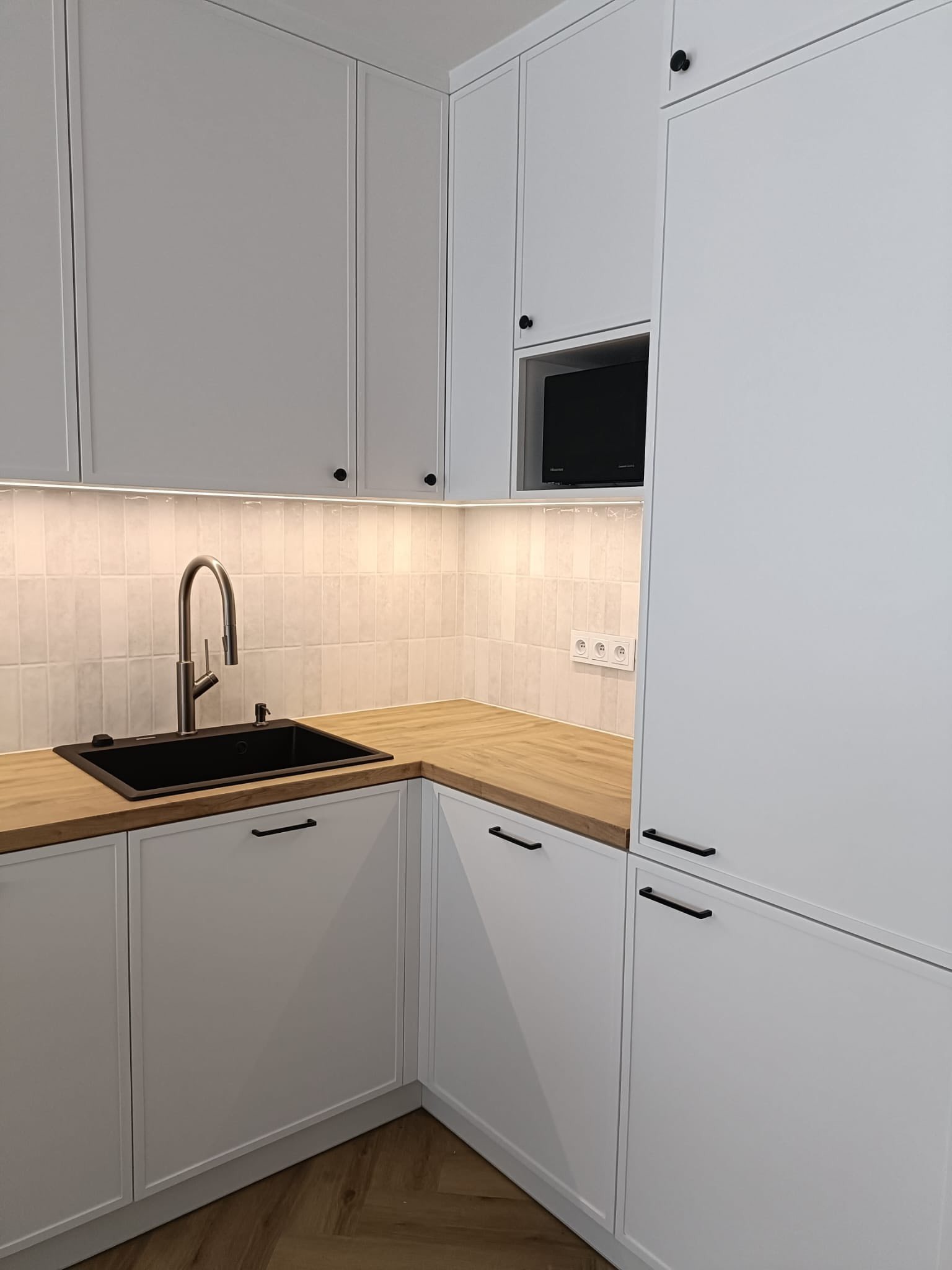Biała kuchnia z frezowanymi frontami i drewnianym blatem. Uwagę przyciąga czarny zlew i bateria. Nad blatem widoczna listwa LED i płytki imitujące cegłę.