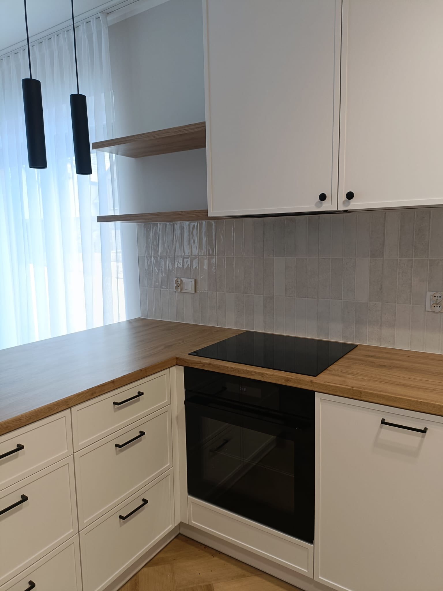 Jasna kuchnia w stylu angielskim: białe frezowane fronty, drewniany blat, czarne, podłużne lampy wiszące. Widoczna płyta indukcyjna i piekarnik.