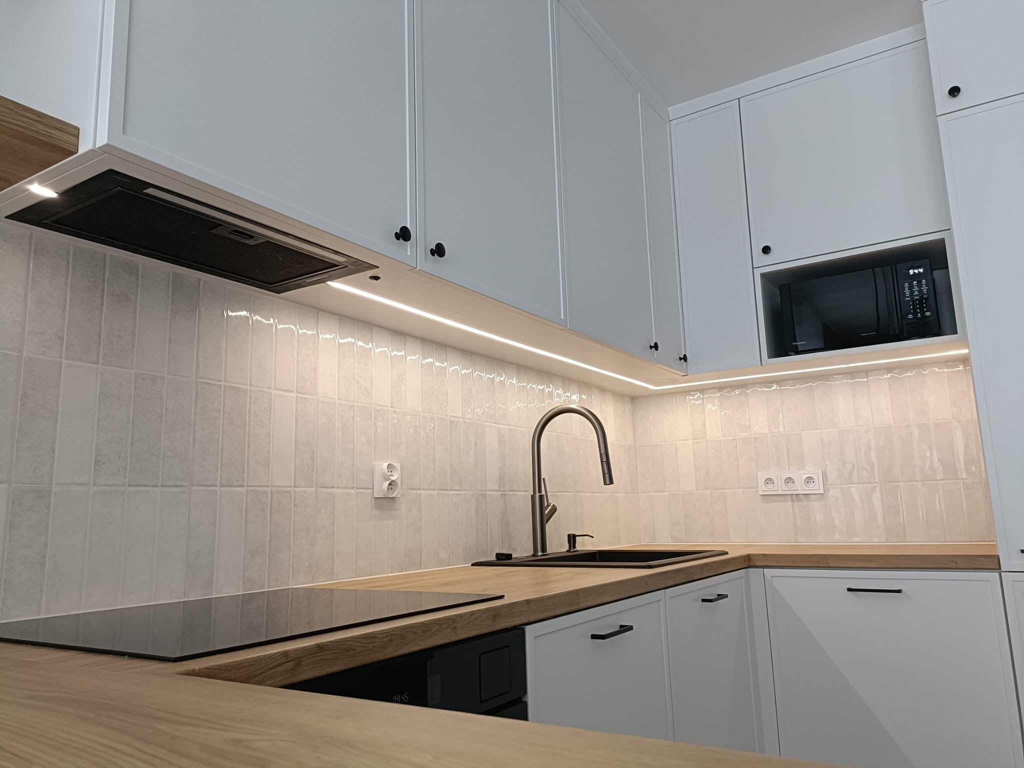 Biała kuchnia z drewnianym blatem i czarnymi akcentami: zlew, bateria, płyta indukcyjna. Frezowane fronty szafek, oświetlenie LED pod szafkami wiszącymi.