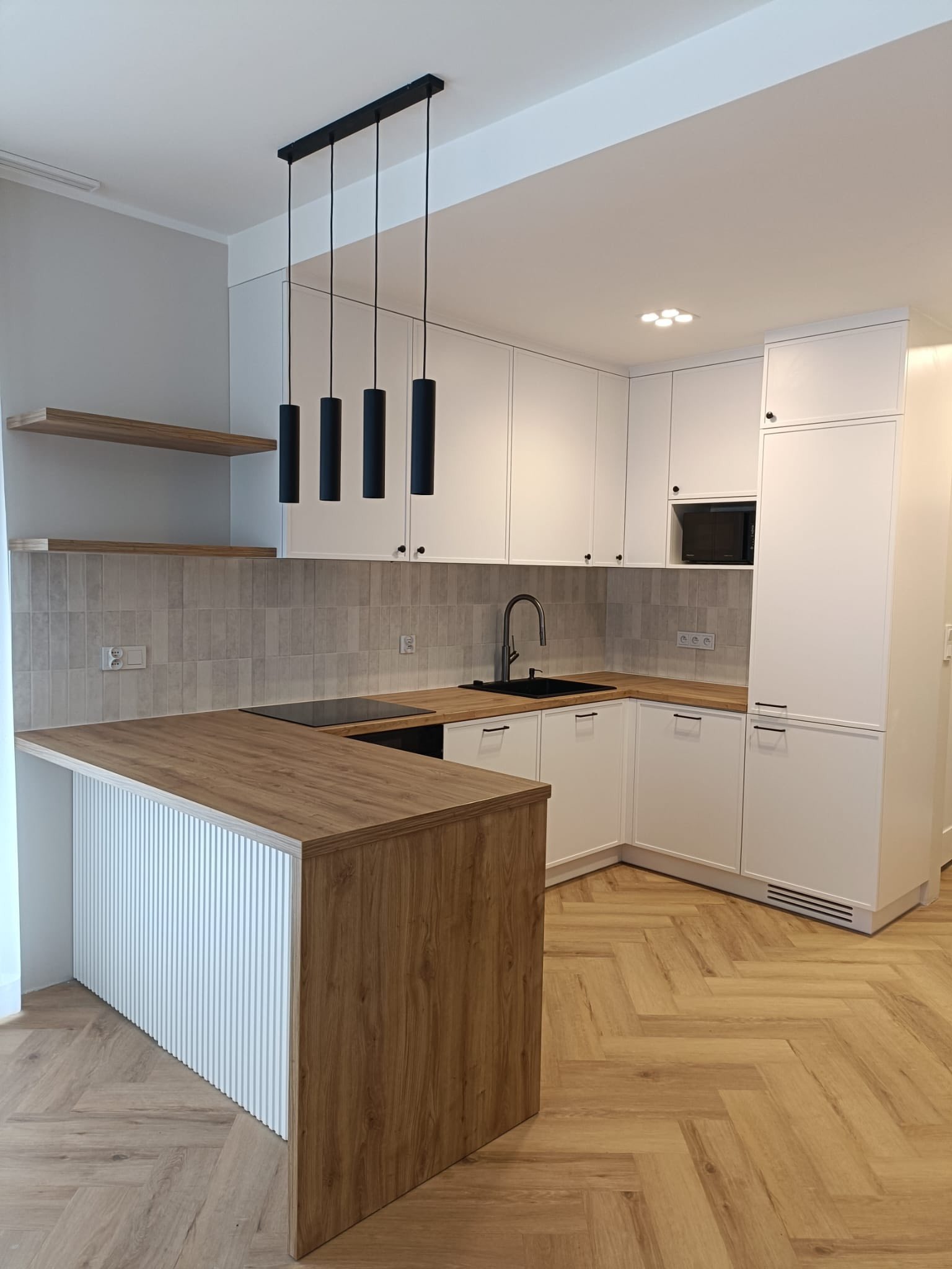 Jasna kuchnia w stylu angielskim: białe, frezowane fronty, drewniany blat i wyspa z ryflowanym wykończeniem. Czarna armatura i lampy dodają kontrastu.