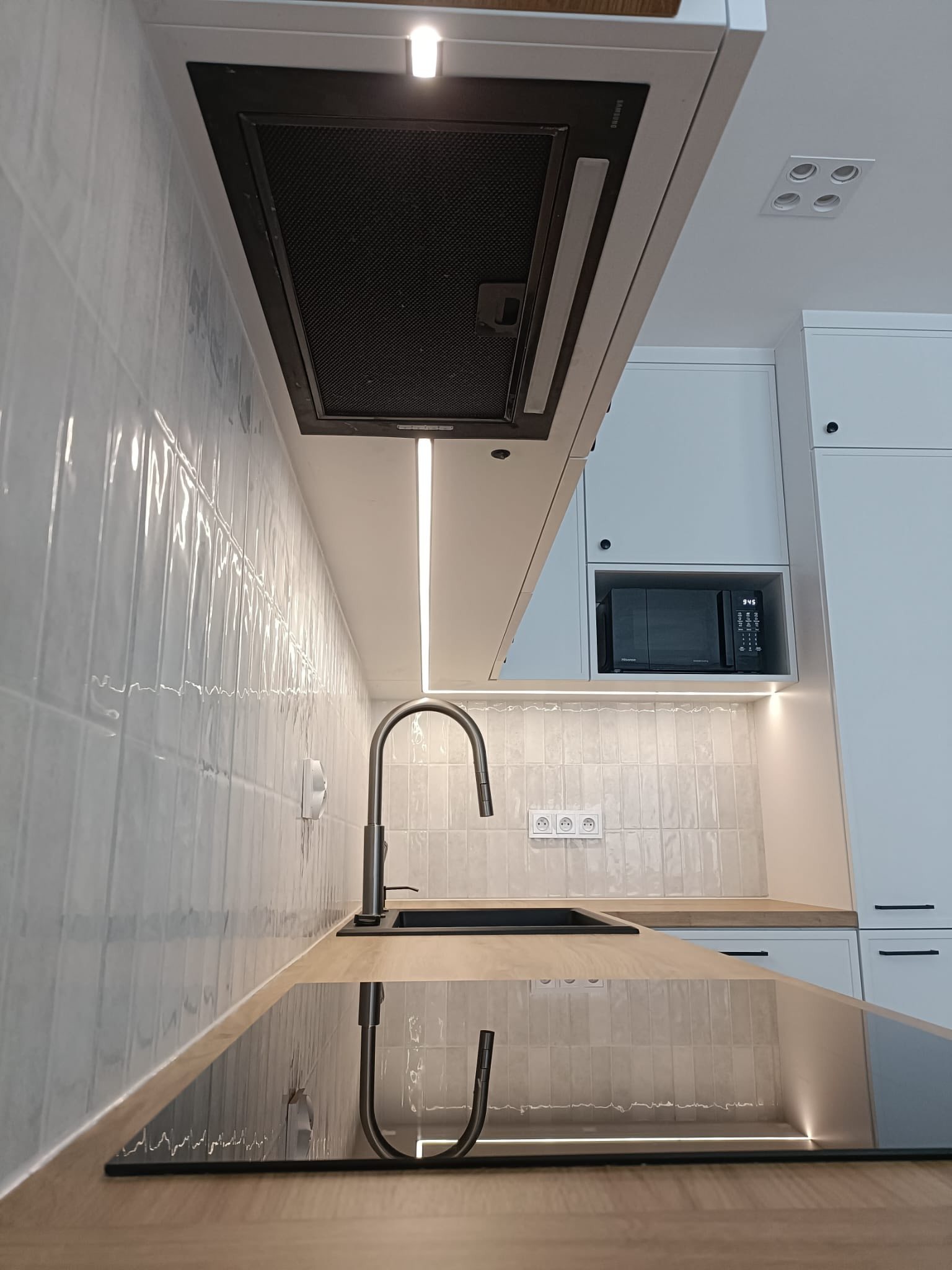 Nowoczesna, jasna kuchnia na wymiar w Krakowie. Białe szafki, drewniany blat, czarny zlew i płyta indukcyjna. Oświetlenie LED podkreśla fakturę płytek.