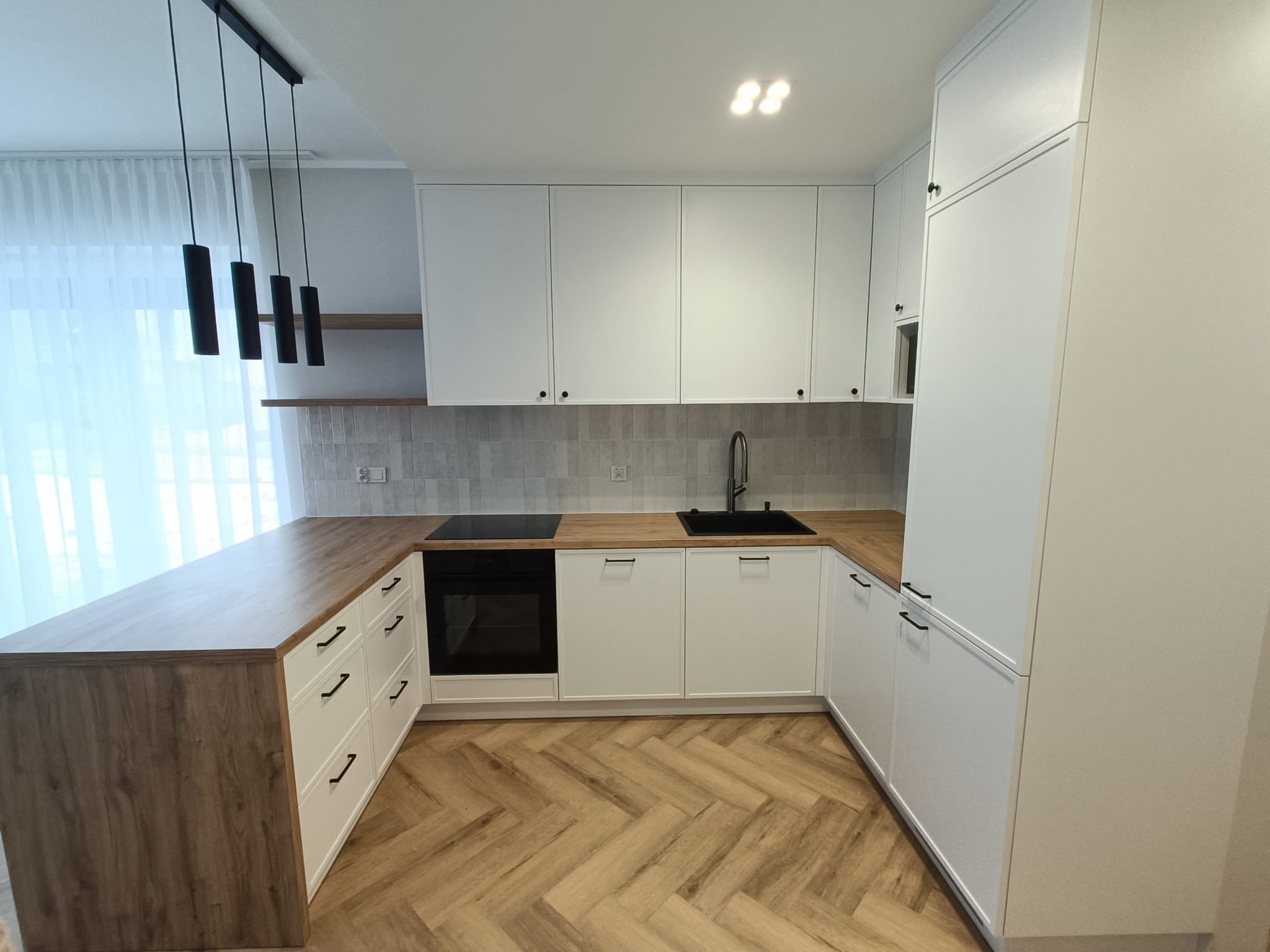 Jasna kuchnia w stylu angielskim z frezowanymi frontami i drewnianym blatem. Cztery czarne lampy wiszące nad blatem. Parkiet ułożony w jodełkę.