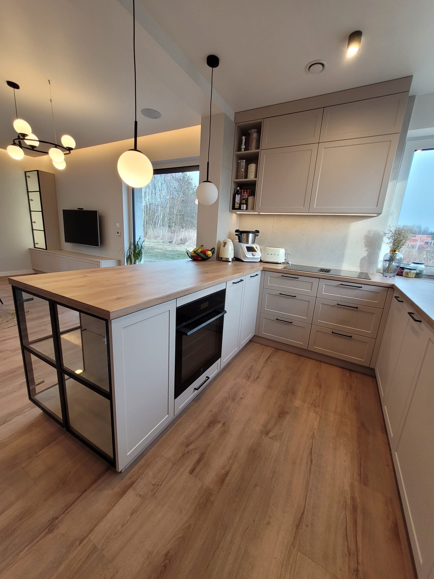 Jasna kuchnia z wyspą i drewnianym blatem. Fronty szafek w odcieniach beżu, czarne, wiszące lampy. Na blacie sprzęt AGD i owoce. Widok na zewnątrz przez duże okno.