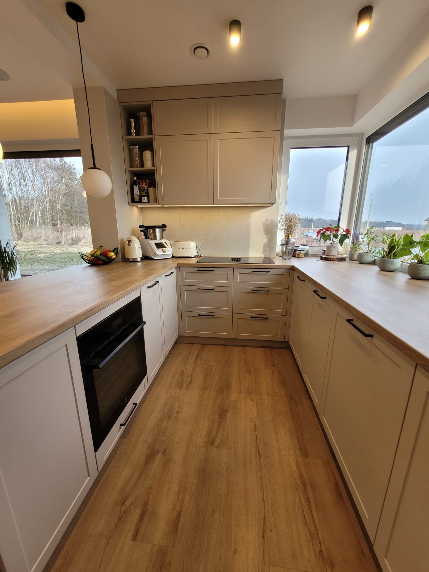 Jasna kuchnia w nowoczesnym stylu z drewnianym blatem, sprzętami SMEG i dużym oknem z widokiem na zewnątrz. Minimalistyczne fronty szafek i drewniana podłoga.