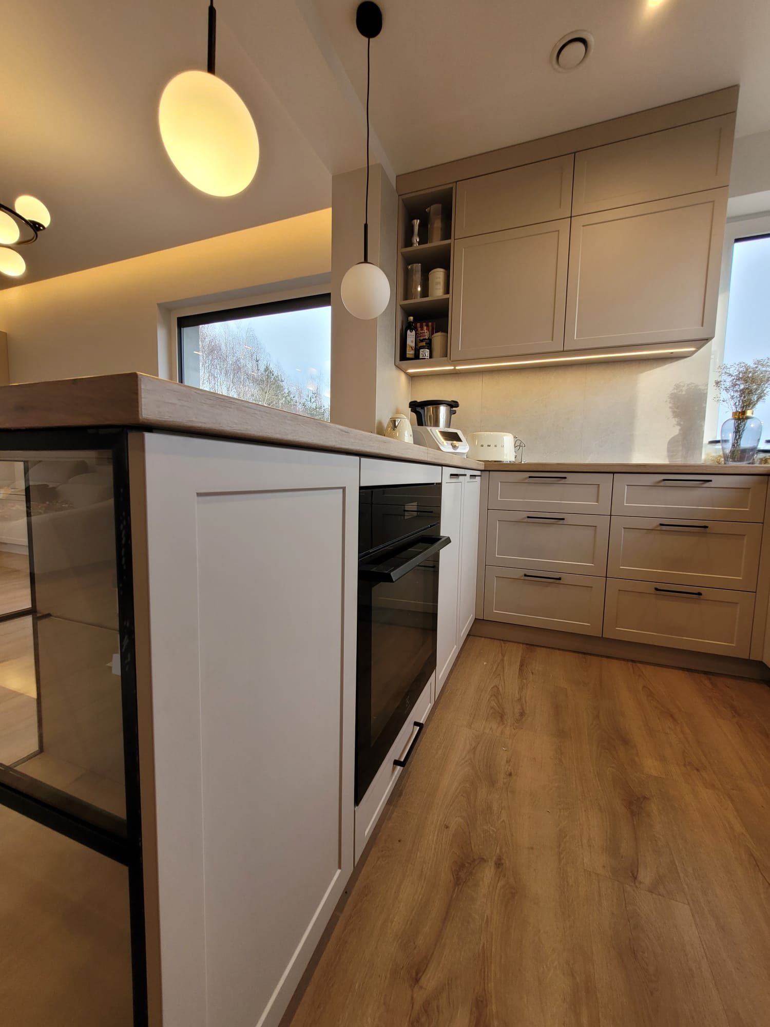 Nowoczesna kuchnia z wyspą, jasne fronty i drewniany blat. Widoczny piekarnik, szafki wiszące z oświetleniem LED. Podłoga drewniana, okno z widokiem na zewnątrz.