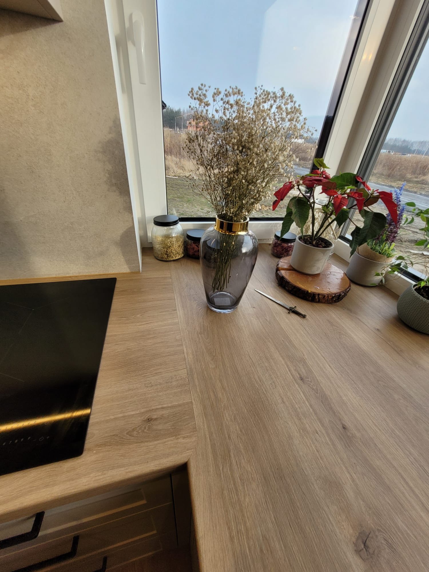 Fragment blatu kuchennego z drewnianym dekorem, na którym stoją wazon z suszem, doniczki z kwiatami i dekoracyjne słoiki. W tle okno z widokiem na zewnątrz.