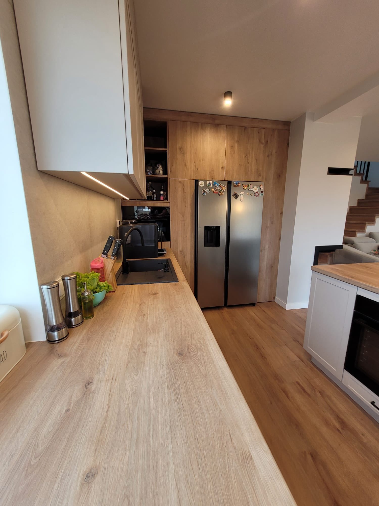 Nowoczesna kuchnia z drewnianym blatem i jasnymi szafkami. Lodówka side-by-side z magnesami. Na blacie przyprawy, zioła i akcesoria kuchenne. Widok na schody w tle.