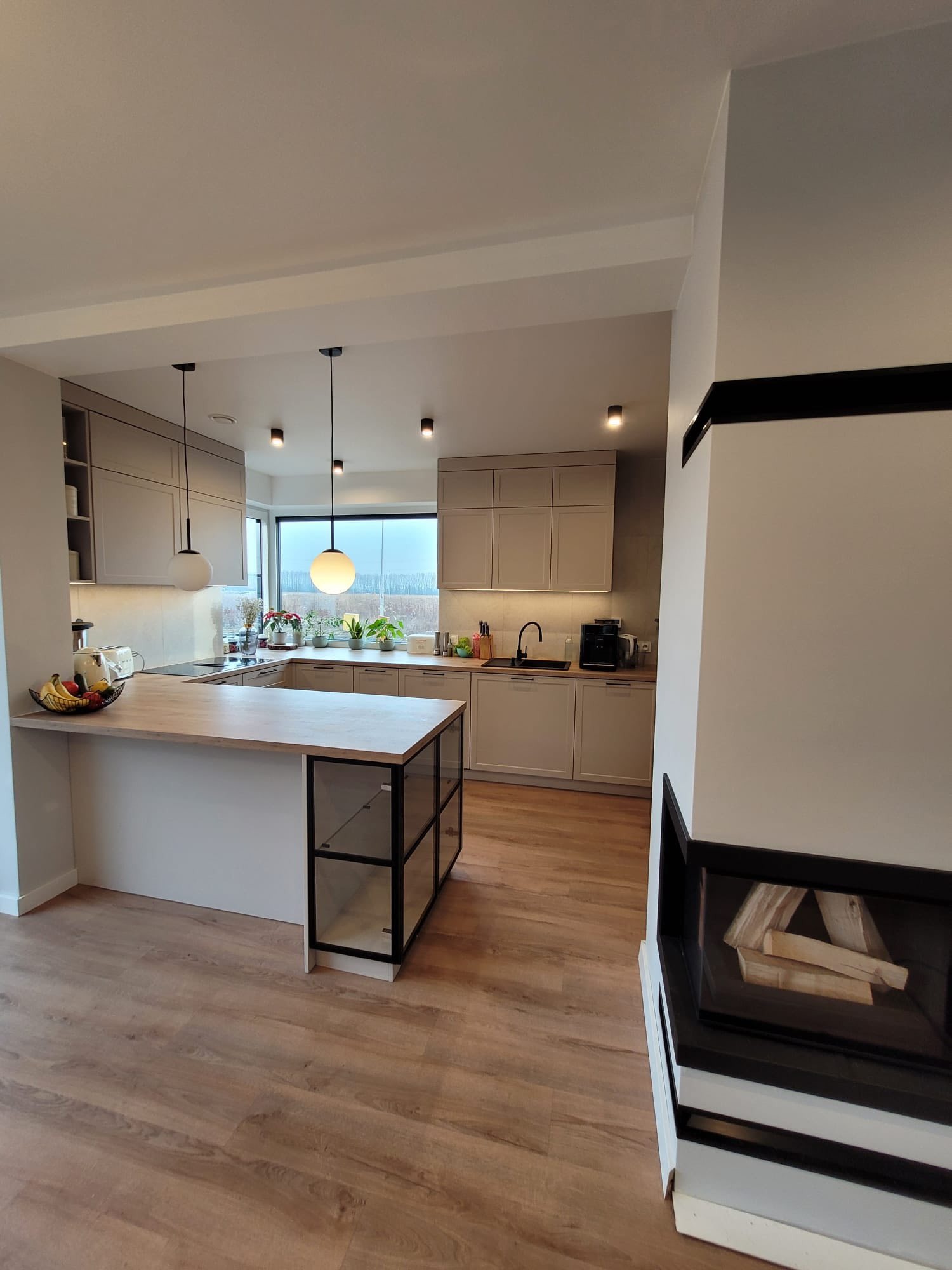 Nowoczesna kuchnia z wyspą i drewnianym blatem. Jasne fronty szafek, czarne lampy wiszące i kominek w salonie tworzą spójną całość. Widok na zieleń za oknem.