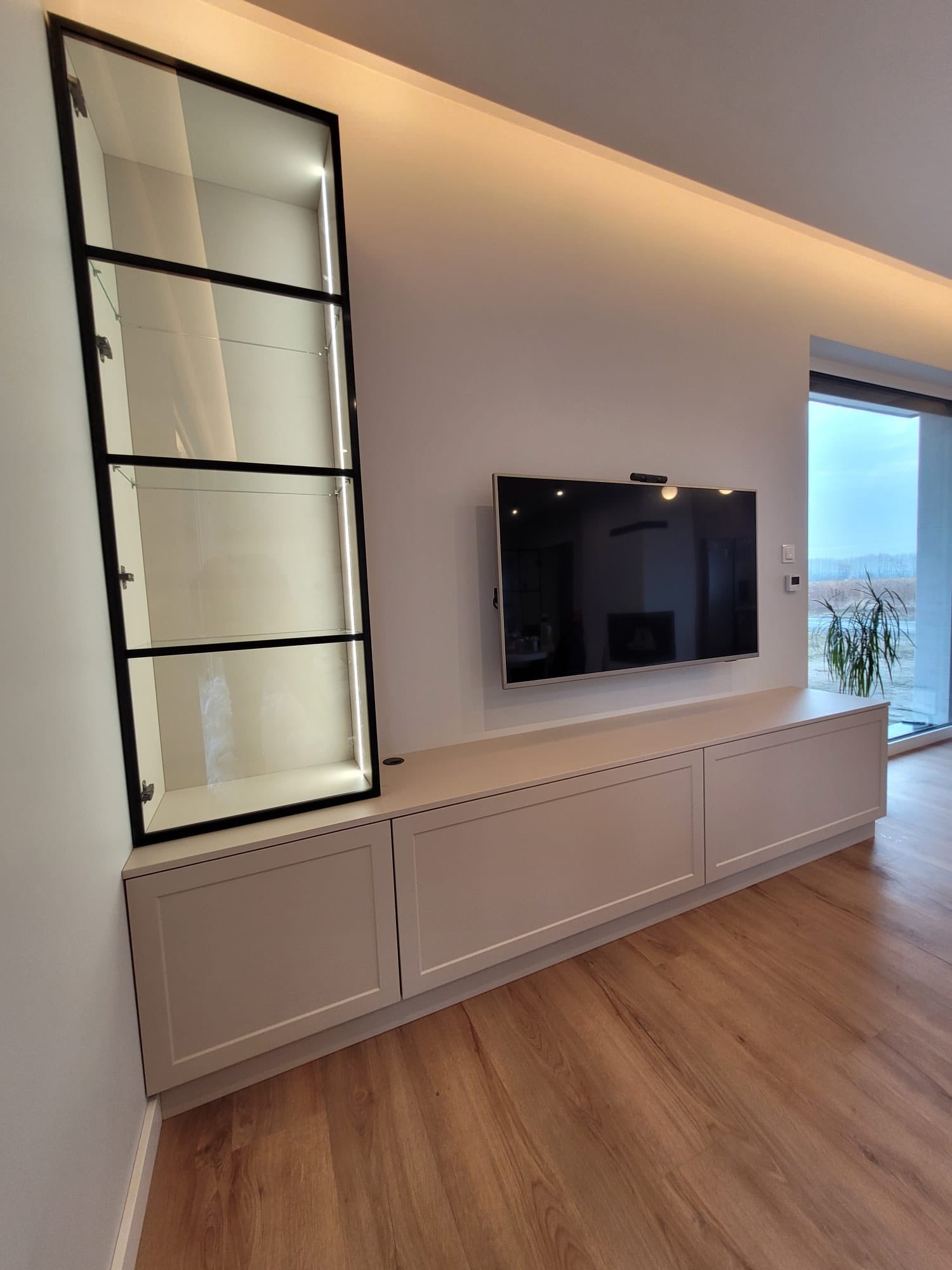 Nowoczesna zabudowa salonu: szafka RTV z szafką wiszącą z czarnymi ramami i oświetleniem LED. Minimalistyczny design, drewniana podłoga, jasne ściany.