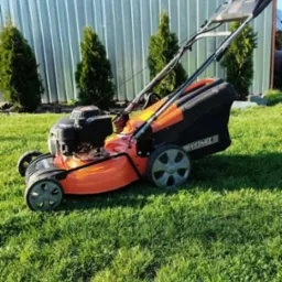 Pomarańczowa kosiarka spalinowa HORTMASZ na trawniku, na tle srebrnego ogrodzenia z blachy trapezowej i trzech małych tuj. Słoneczny dzień.